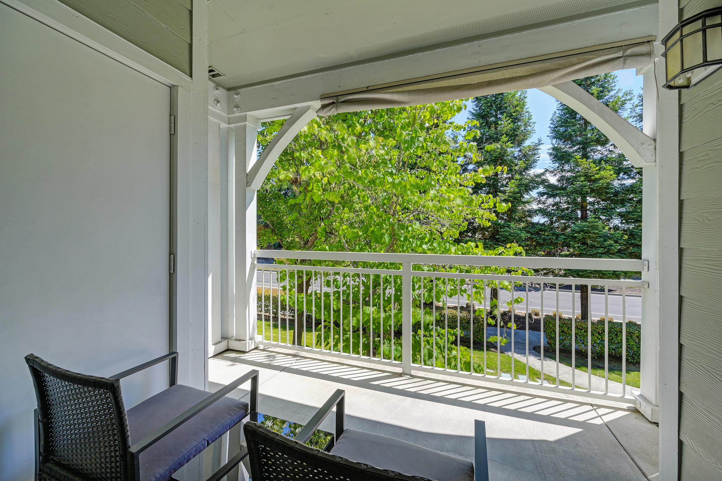 A serene balcony view featuring two black outdoor chairs and a small table, surrounded by green trees and a well-maintained garden. Sunlight filters through the leaves, creating a peaceful atmosphere and inviting outdoor space.