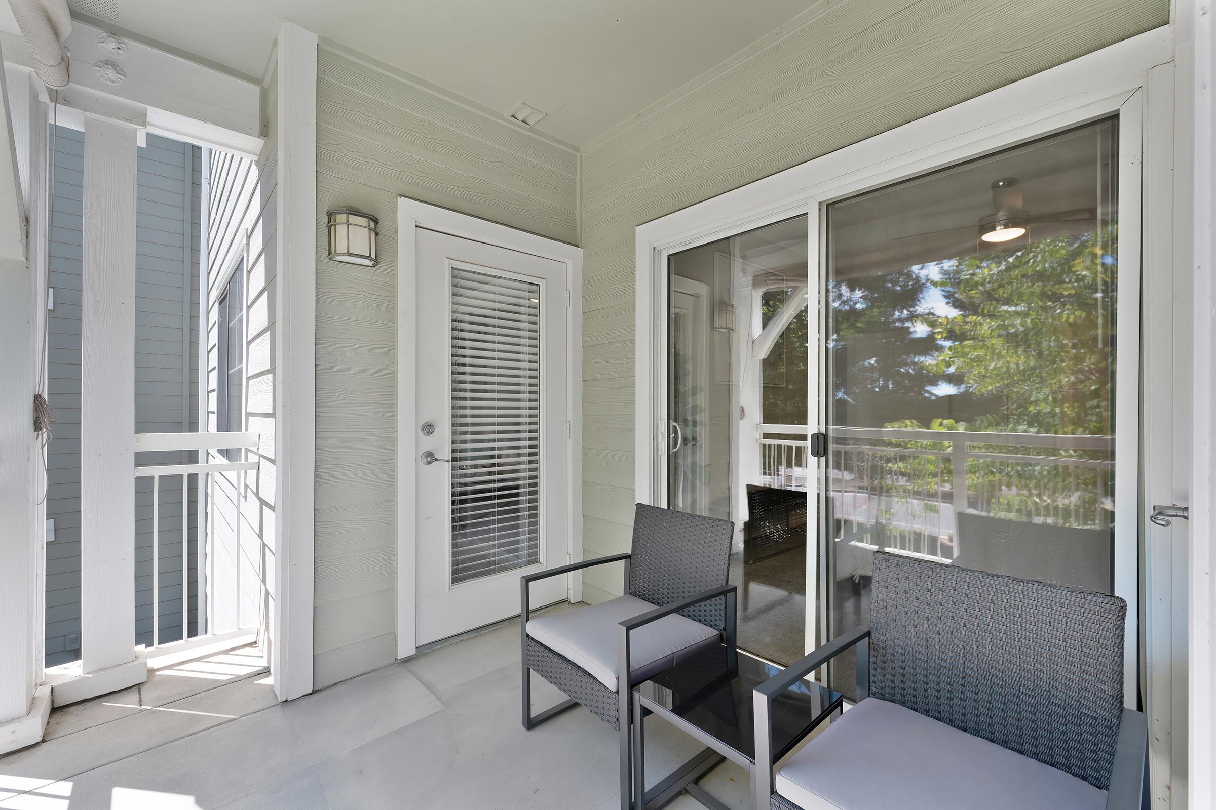 A cozy balcony area featuring two modern wicker chairs with light-colored cushions, a glass door leading to an interior space, and a wall lamp. The walls are painted in a soft green hue, and there’s a view of greenery outside the glass door, creating a tranquil atmosphere.