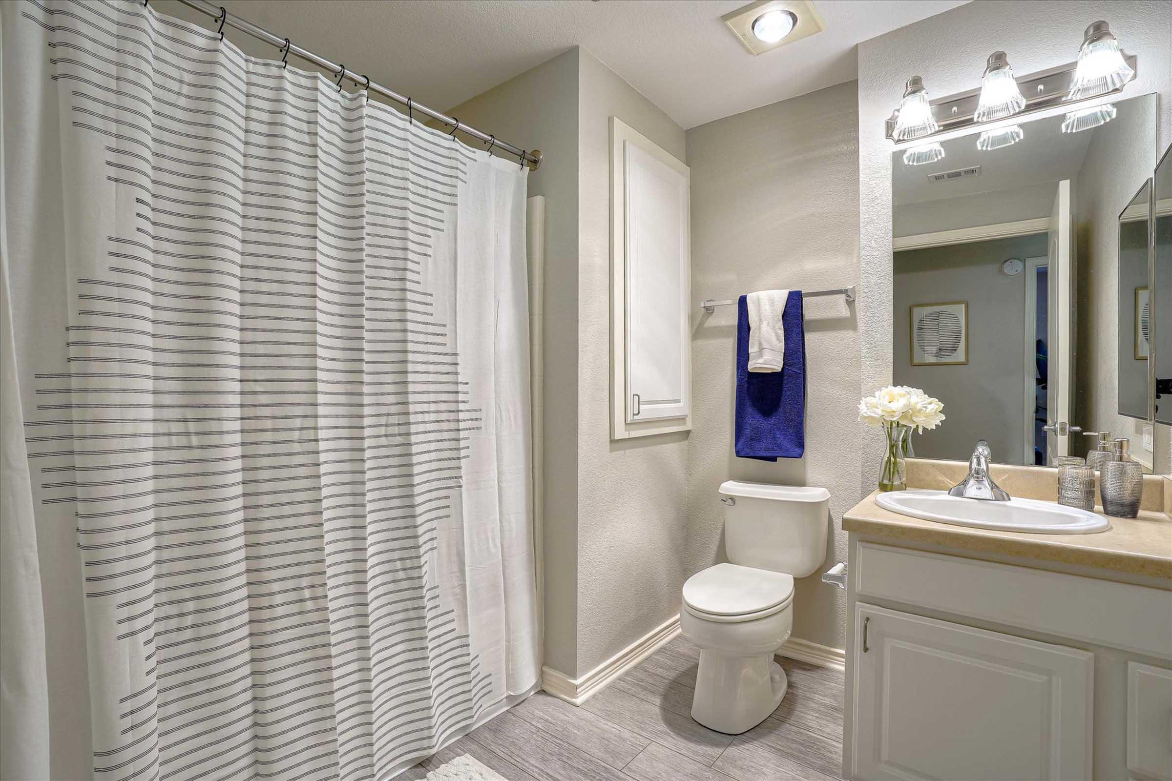 A modern bathroom featuring a white shower curtain with gray stripes, a clean toilet, and a sink with a countertop. The wall is painted a light color, and there's a towel hanging on a rack. A vase of flowers adds a decorative touch, and there's a mirror reflecting the space.