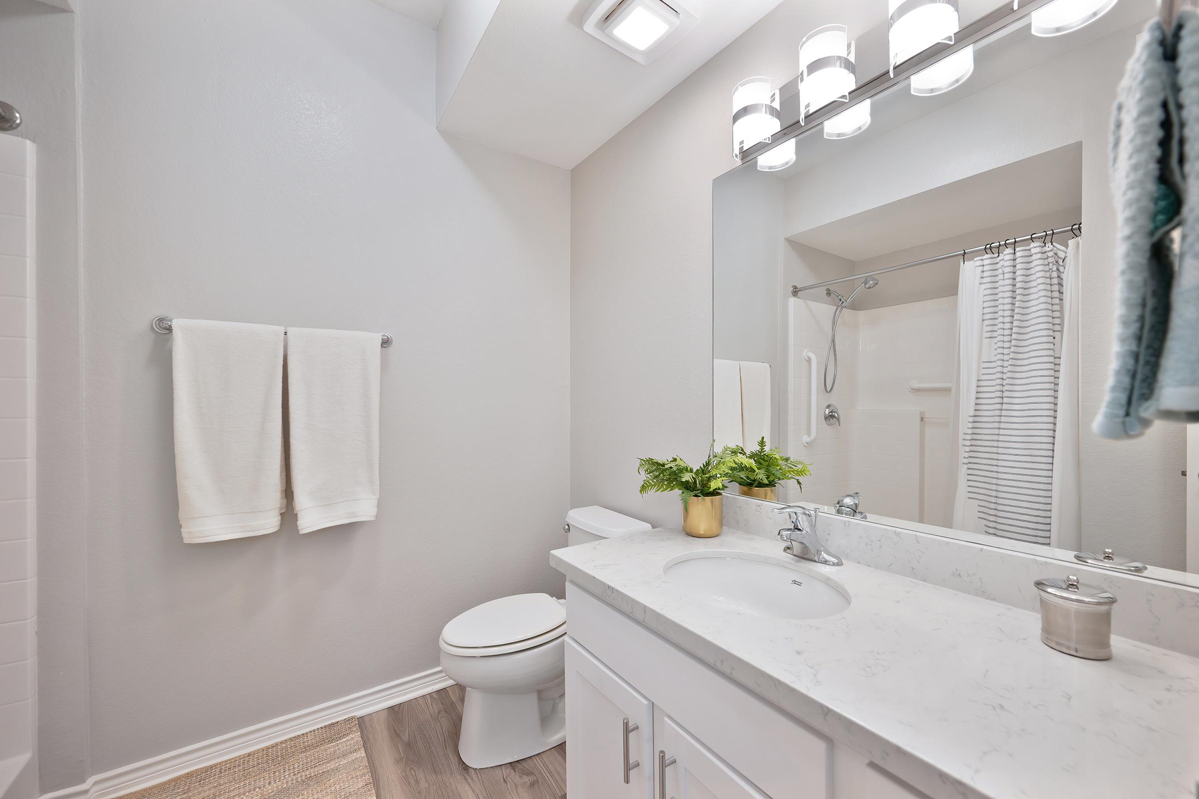 A modern bathroom featuring a white sink with a marble countertop, a large mirror, and a stylish light fixture. White towels are hung on a towel rack, and a small plant sits in a gold pot. The shower area is partially visible in the background, along with light-colored flooring and neutral wall colors.