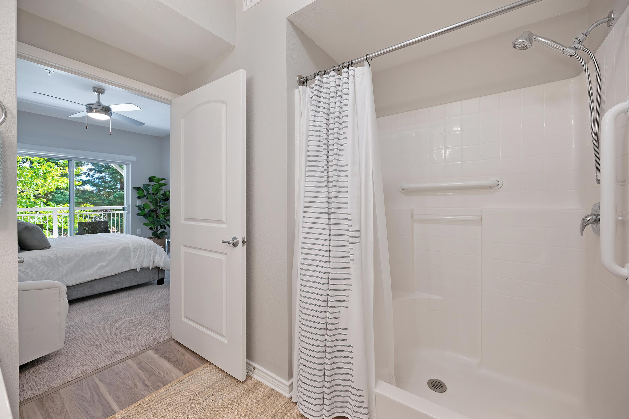 A bathroom with a shower featuring a white curtain, looking out towards a bedroom with a ceiling fan and large windows. The room beyond has a bed and greenery visible outside, creating a bright and airy atmosphere. The bathroom is well-lit with neutral-colored walls and clean lines.