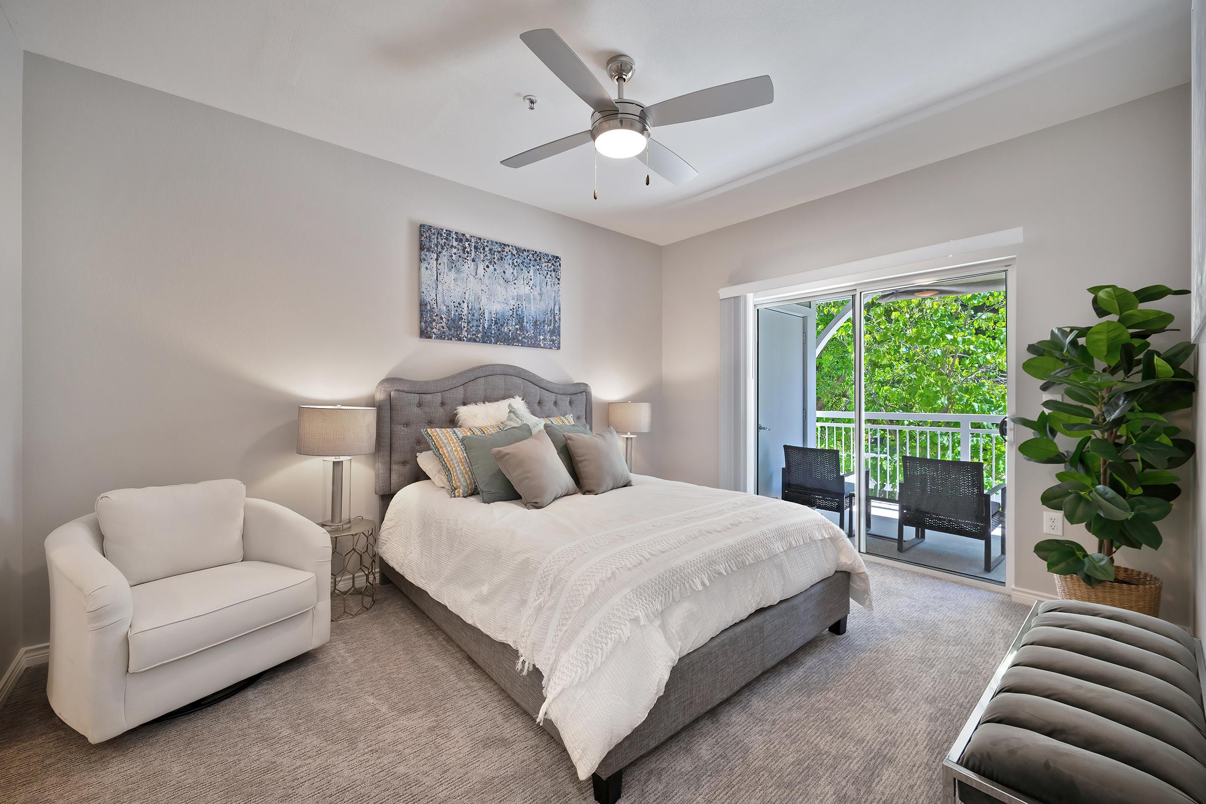 A modern bedroom featuring a queen-sized bed with a light gray upholstered headboard and fluffy white bedding. There are two bedside lamps on either side, a small white armchair, and a decorative plant. A sliding glass door leads to a balcony with two black chairs and green foliage visible outside.