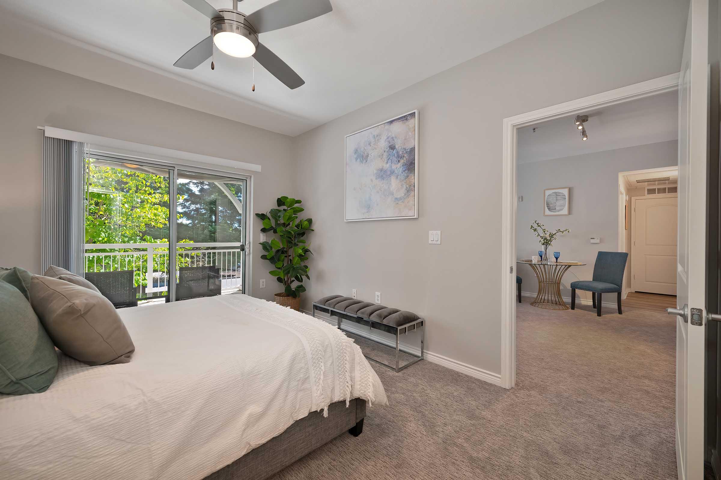 A cozy bedroom featuring a large bed with gray and teal pillows, a ceiling fan, and a window with outdoor views. A potted plant stands beside the bed, and a stylish bench sits at the foot. The room leads to a hallway with a small table and chairs, enhancing the modern, inviting atmosphere.