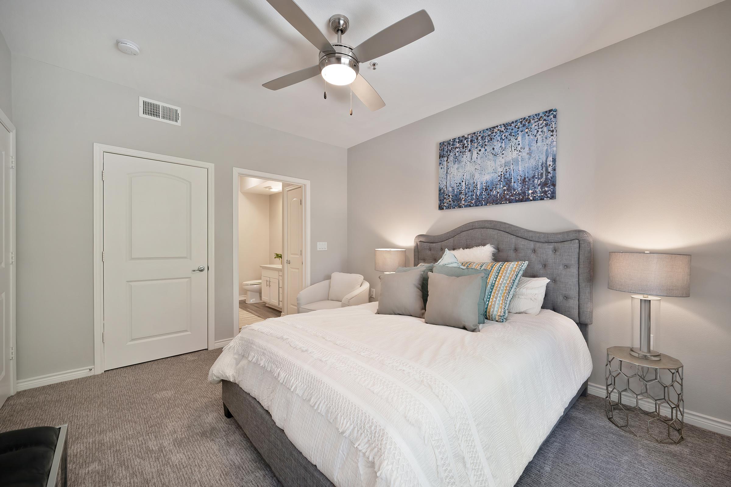 A stylish bedroom featuring a gray upholstered bed with white bedding and decorative pillows. Two bedside lamps provide soft lighting. A colorful piece of art hangs above the bed, and a door leads to an adjacent bathroom. The room has light gray walls and carpet, creating a cozy and inviting atmosphere.