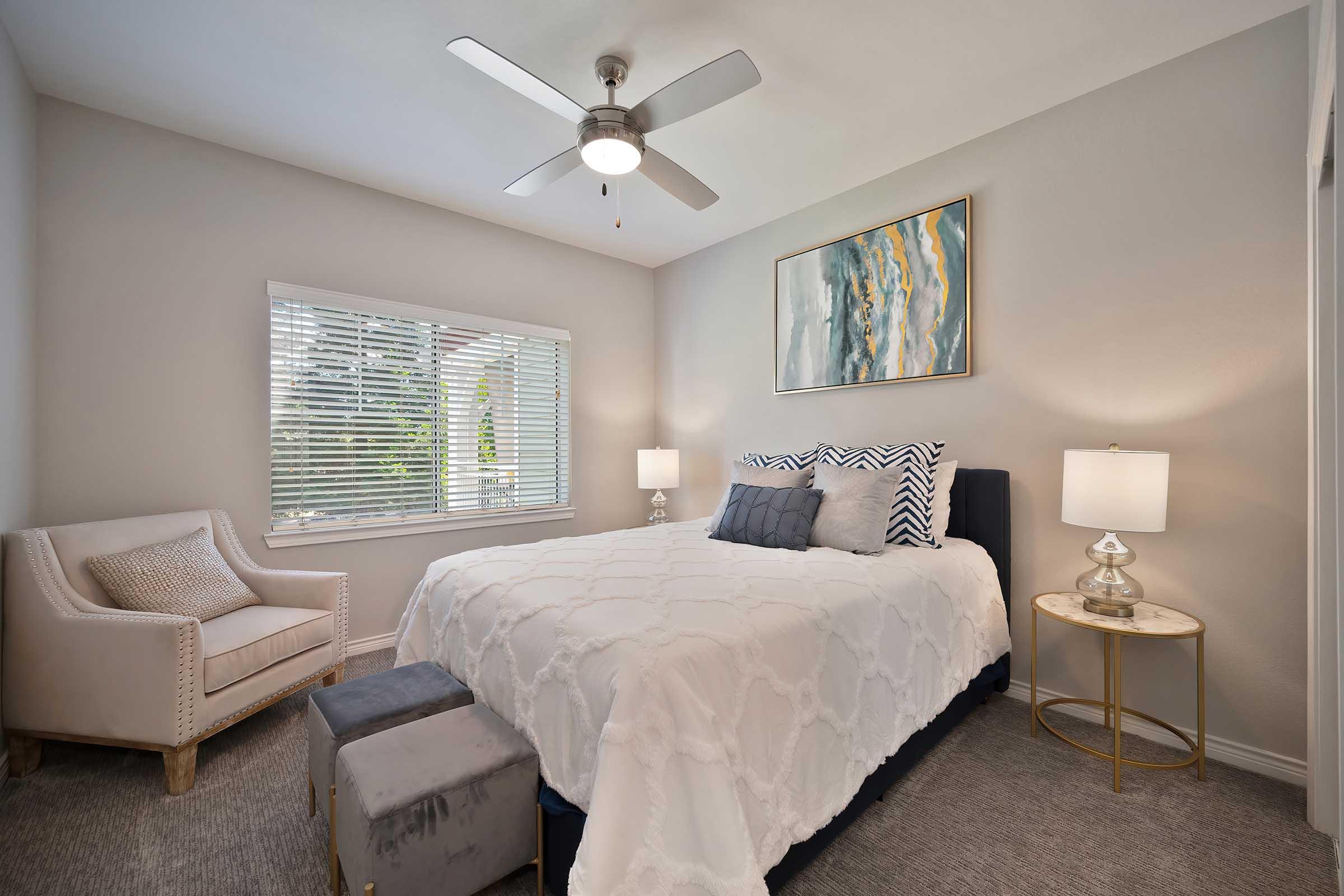 A cozy bedroom featuring a queen-sized bed with a white quilt, decorative pillows, and a light gray armchair. The room is well-lit by a ceiling fan and has two bedside lamps. A window with white blinds allows natural light, and a piece of abstract art hangs above the bed, adding a touch of modern decor.