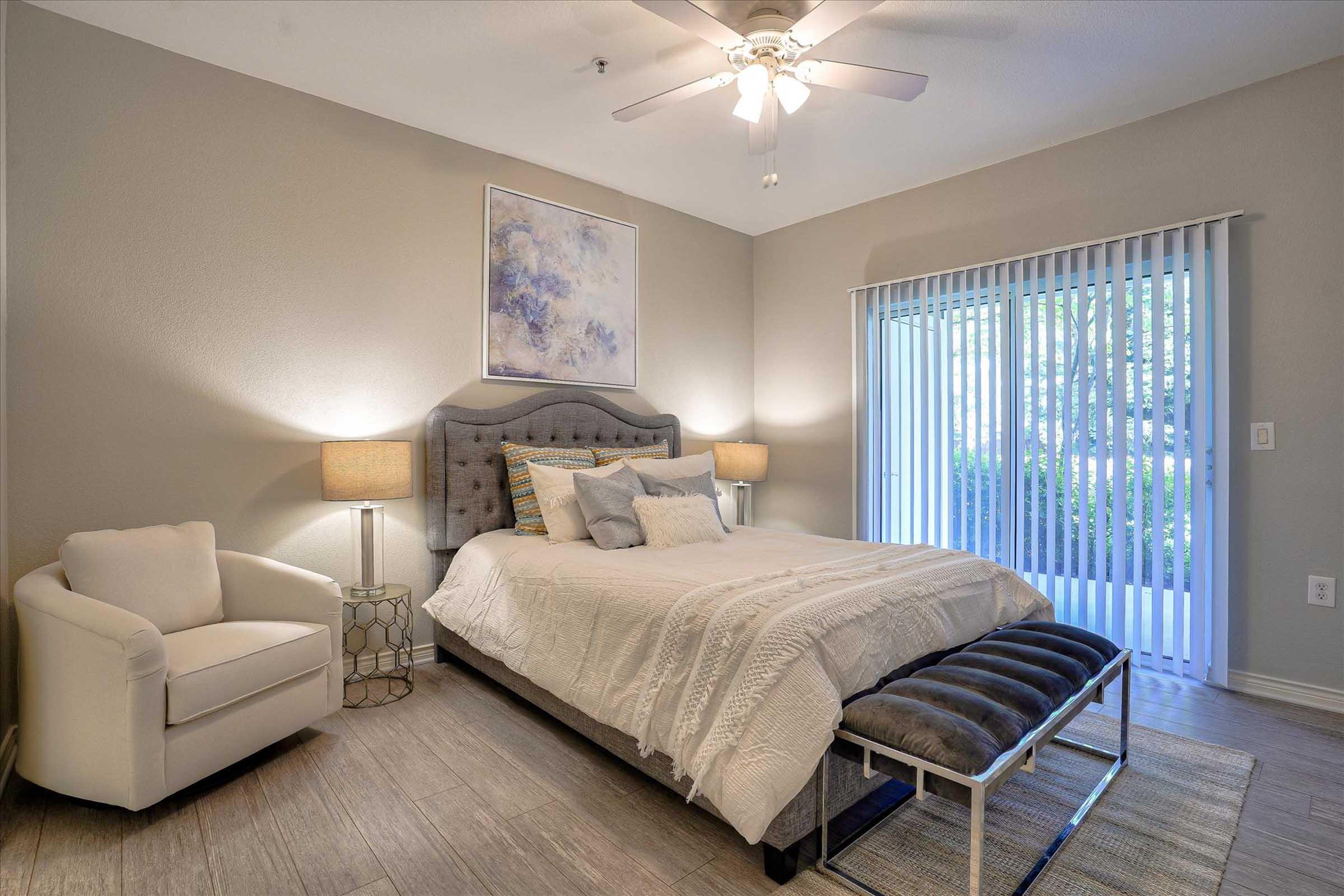 A cozy bedroom featuring a queen-sized bed with a grey tufted headboard and white bedding. A light-colored armchair sits beside the bed, and two bedside lamps provide soft lighting. There's a bench at the foot of the bed, and large sliding glass doors lead to a patio area with natural light filtering in.