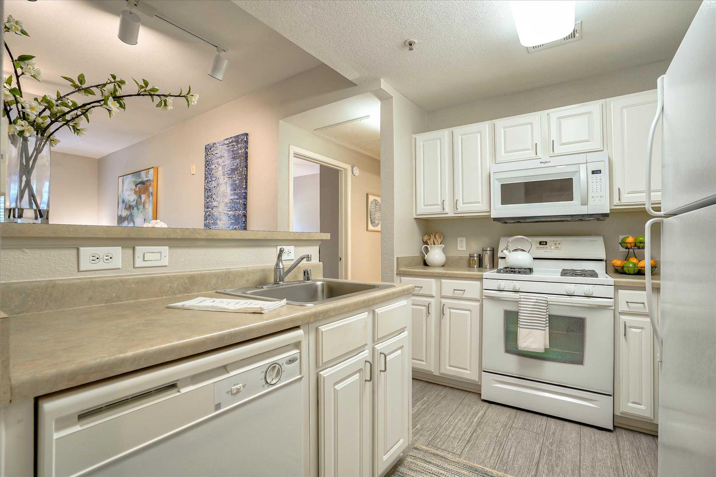 A modern kitchen featuring white cabinetry, a double sink, a dishwasher, a stove, and a microwave. The countertops are light-colored, and a small decorative plant is visible. The space is well-lit and organized, with some kitchen utensils and fruits on display.