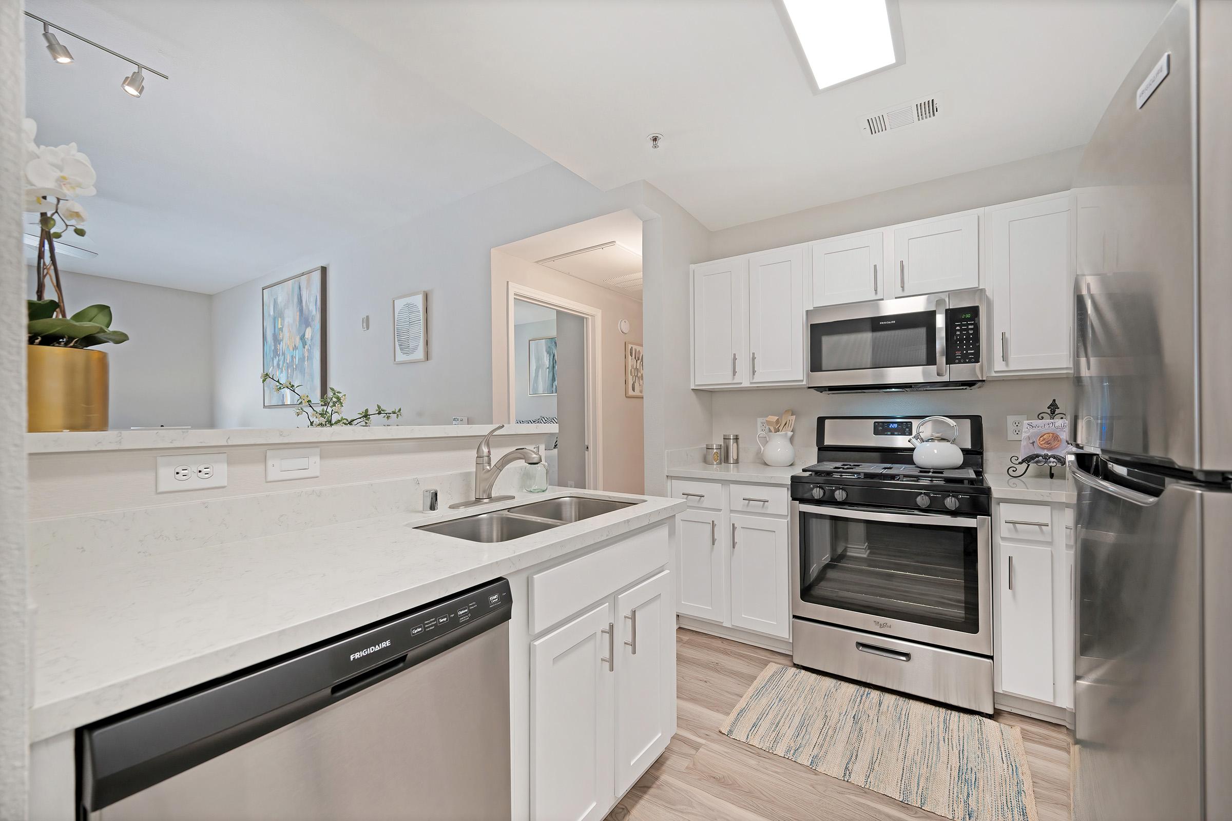 Modern kitchen featuring white cabinetry, stainless steel appliances including an oven and refrigerator, a dishwasher, and a gray countertop. The space is well-lit with a ceiling light, and decorative elements like a potted plant and artwork can be seen in the background. Hardwood-style flooring completes the look.