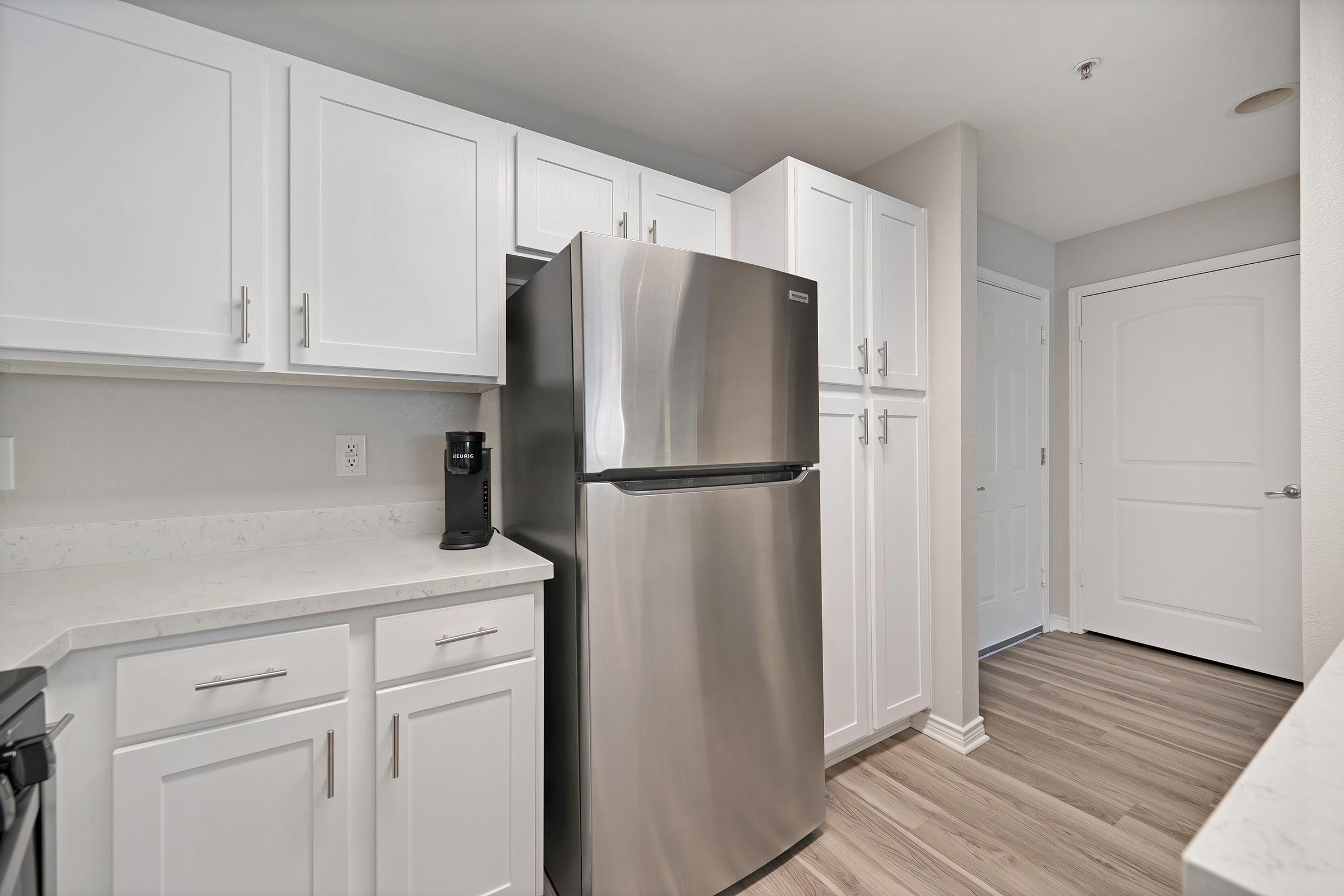 A modern kitchen featuring white cabinetry, a stainless steel refrigerator, and light-colored countertops. The floor is a light wood laminate, and the space is well-lit with minimal decoration, showcasing a clean and contemporary design. A doorway leads to another room.