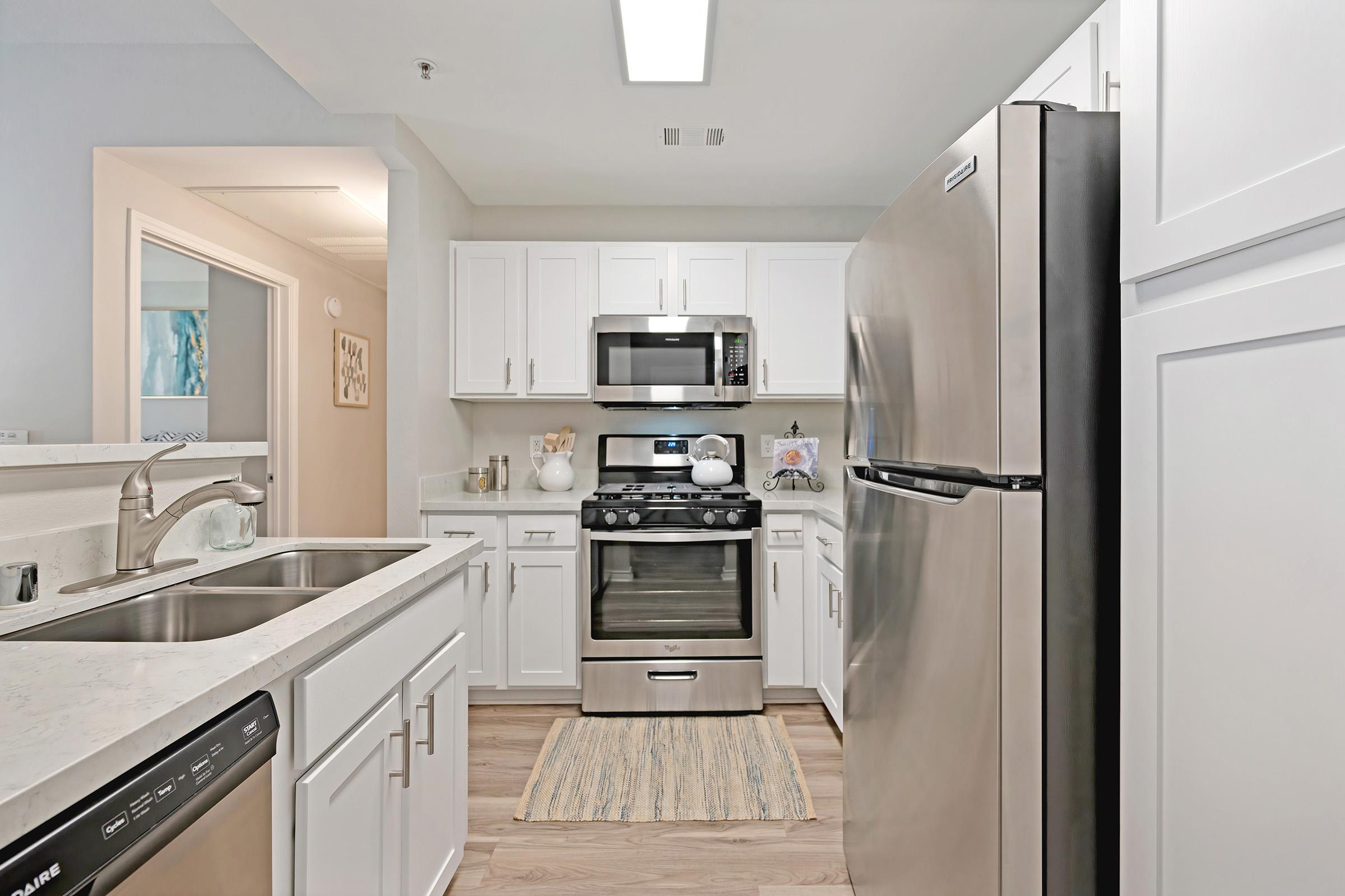 Modern kitchen featuring stainless steel appliances, including a refrigerator, oven, and microwave. The white cabinetry complements the light-colored countertops, while a rug adds a cozy touch to the space. Natural light floods the room, creating a bright and inviting atmosphere.