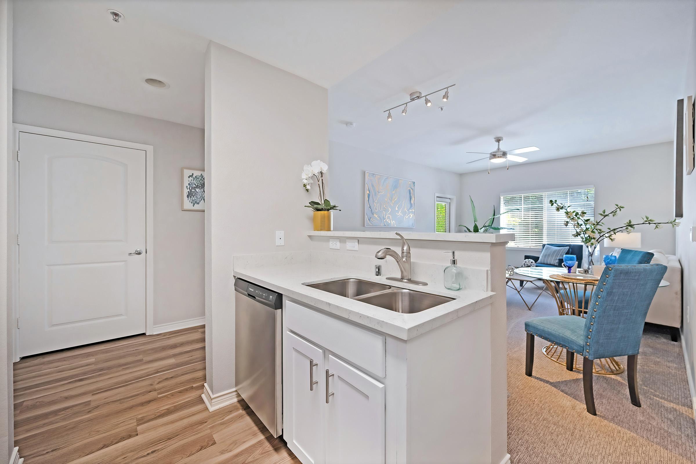 Modern kitchen with a double sink and dishwasher, featuring white countertops. In the background, a cozy living area with a blue chair, a round dining table, and large windows allowing natural light. The walls are light-colored, and decor includes plants and artwork.