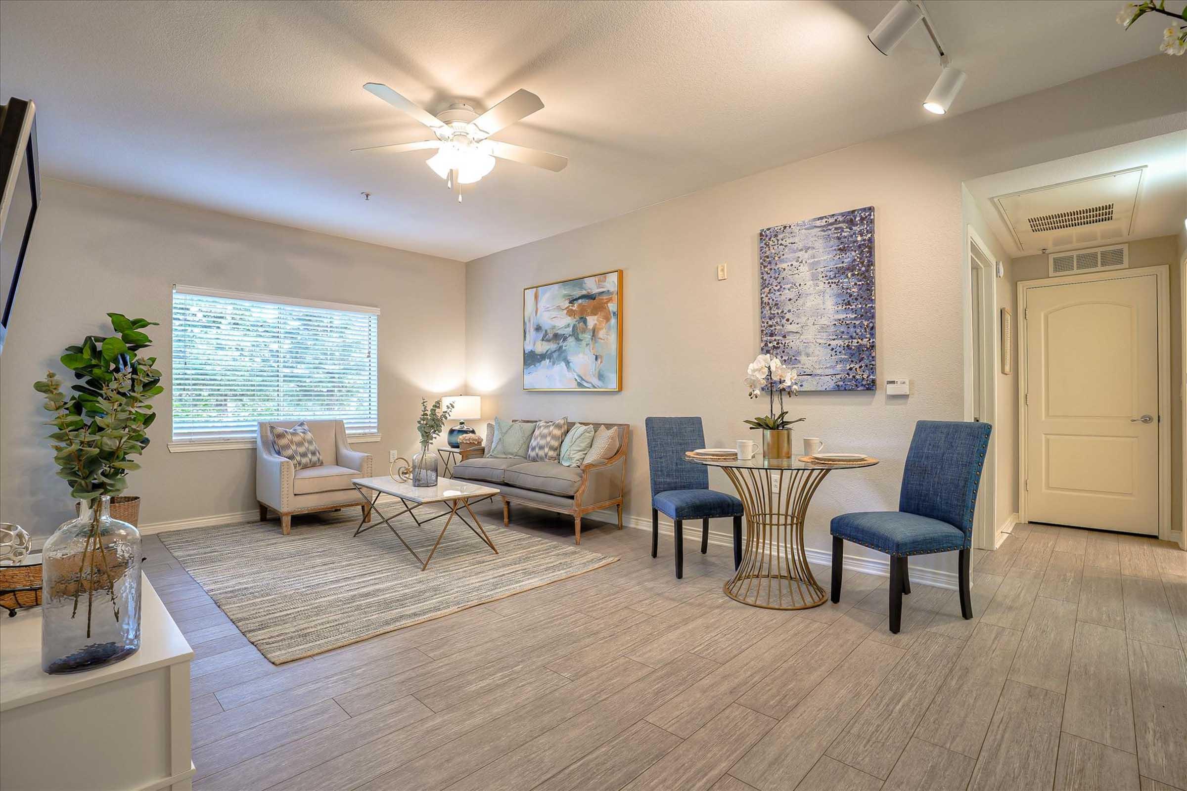 A well-decorated living room featuring a light-colored sofa, a coffee table, and two blue chairs placed at a small dining table. The space is brightened by natural light coming through a window and is adorned with modern artwork and decorative plants, creating a cozy and inviting atmosphere.