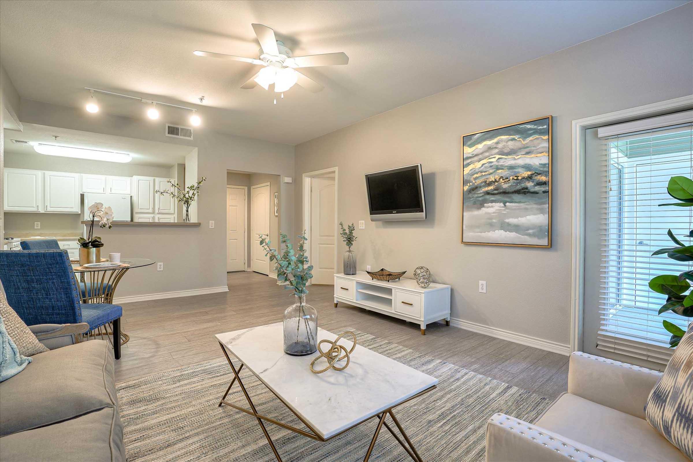 A modern living room featuring a light-colored sofa, a coffee table with a marble top, and decorative items. There's a flat-screen TV mounted on the wall, a large piece of abstract art, and a window with blinds. Potted plants add a touch of greenery, and the space is illuminated by a ceiling fan.