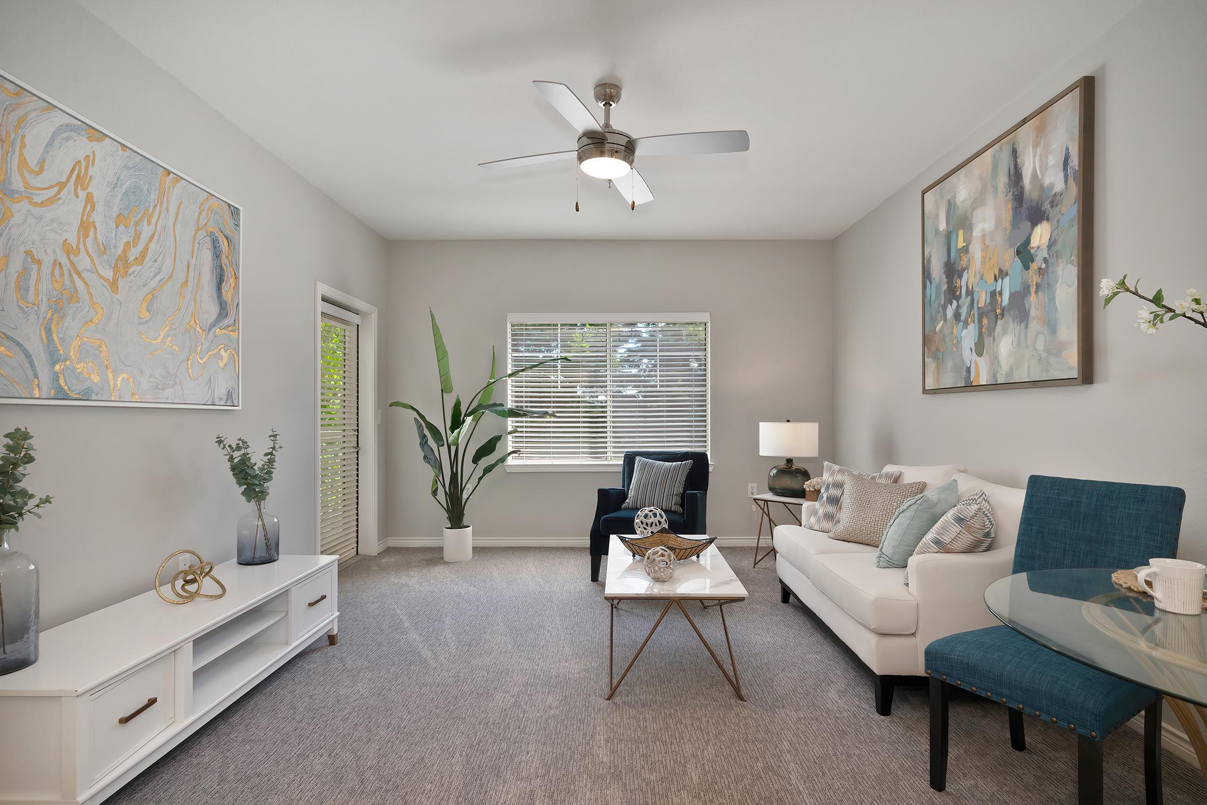 A bright, modern living room featuring a white sofa, a blue accent chair, and a glass coffee table. A large artwork hangs on the wall, complemented by decorative plants. The space has large windows allowing natural light, with a light carpet and minimalist decor, creating an inviting atmosphere.