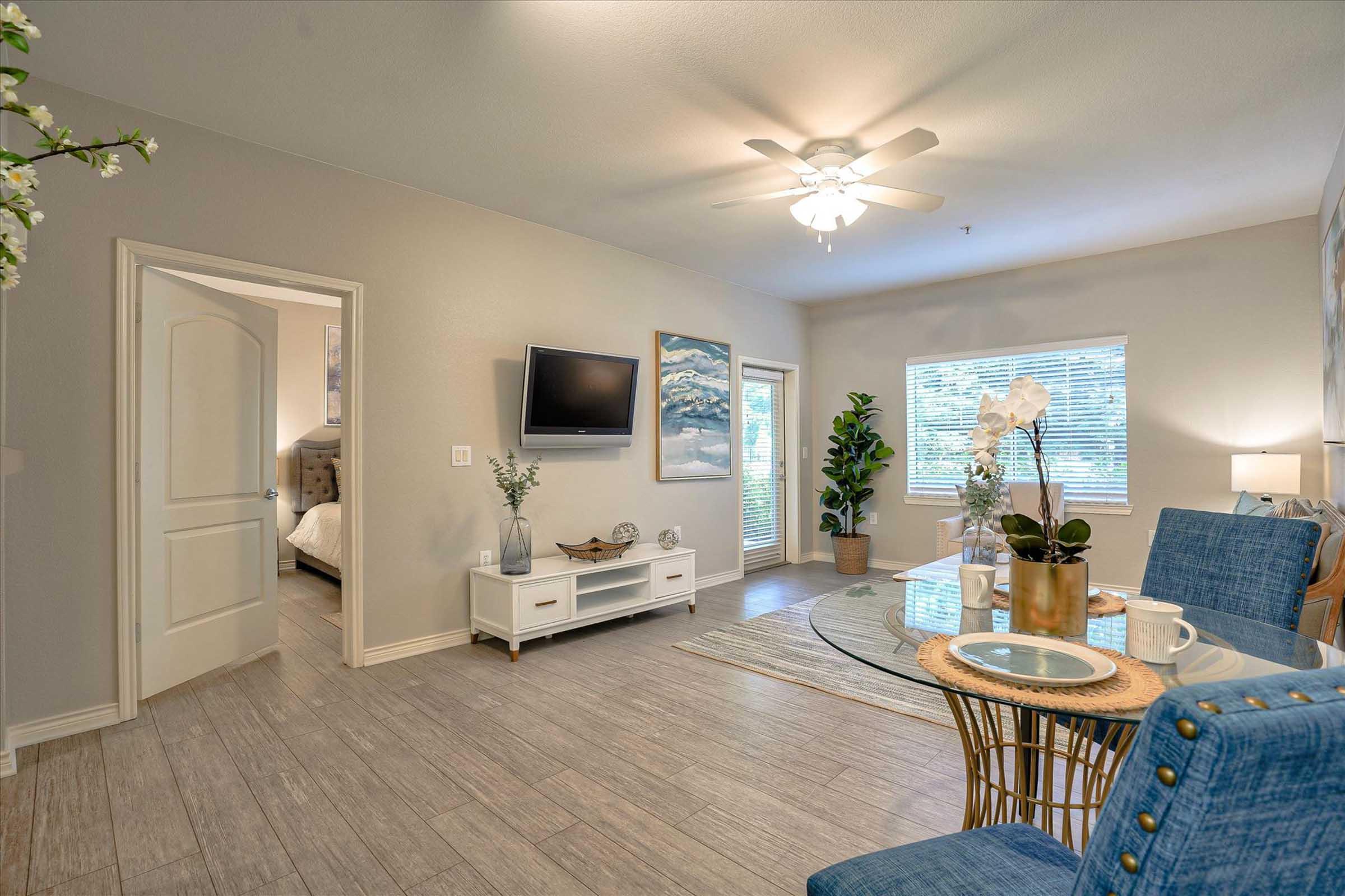 A modern living space featuring a light-colored wall, a ceiling fan, and a mounted TV. A round dining table with blue chairs sits in the foreground, surrounded by decorative plants and a vase with flowers. A doorway leads to another room, and natural light filters in through a window.