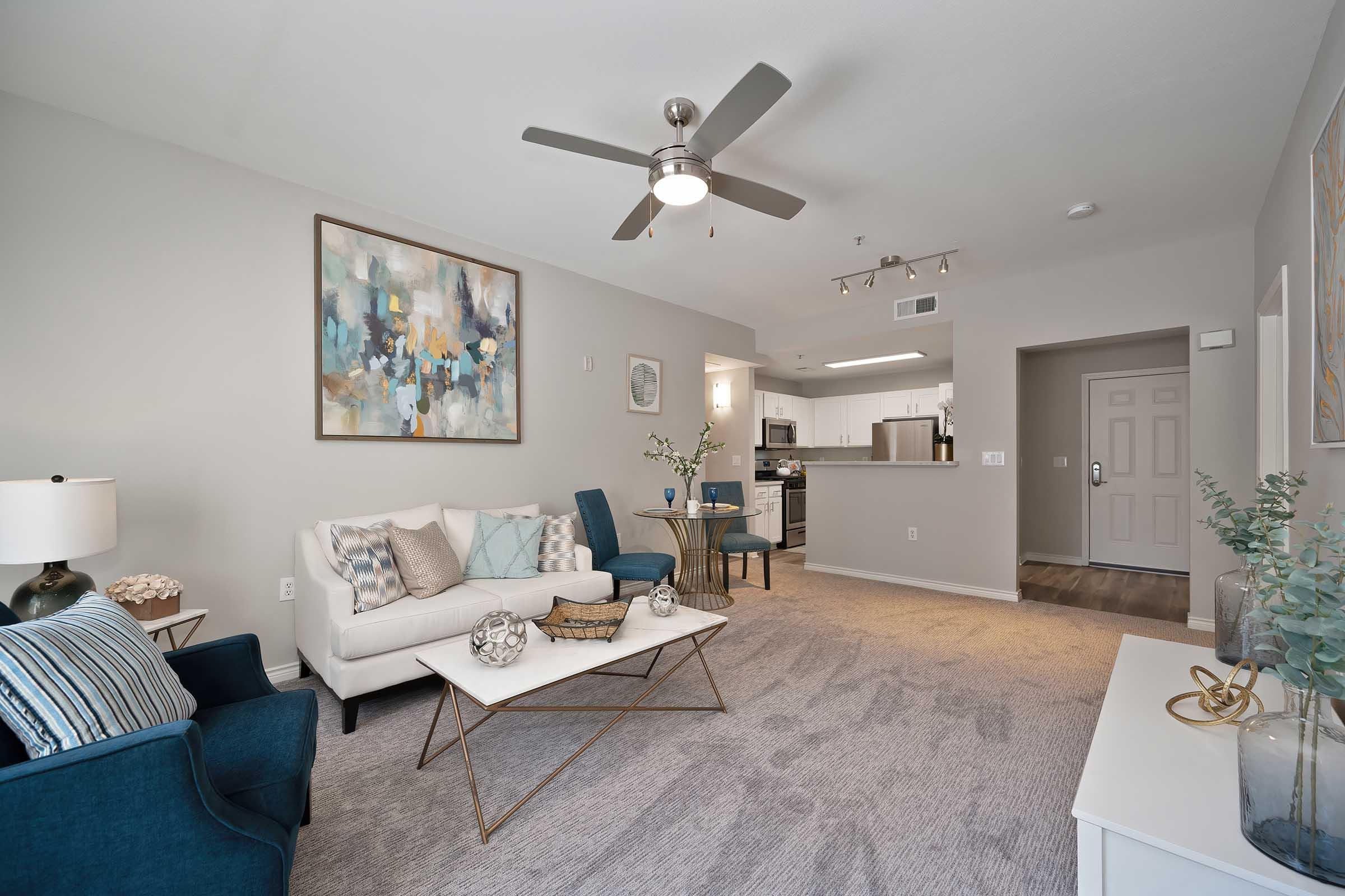 A modern living room featuring a light gray color scheme. There’s a white sofa with decorative pillows, a stylish coffee table, and two blue chairs. A ceiling fan hangs above, and artwork is displayed on the walls. The kitchen is visible in the background, along with a front door leading outside.