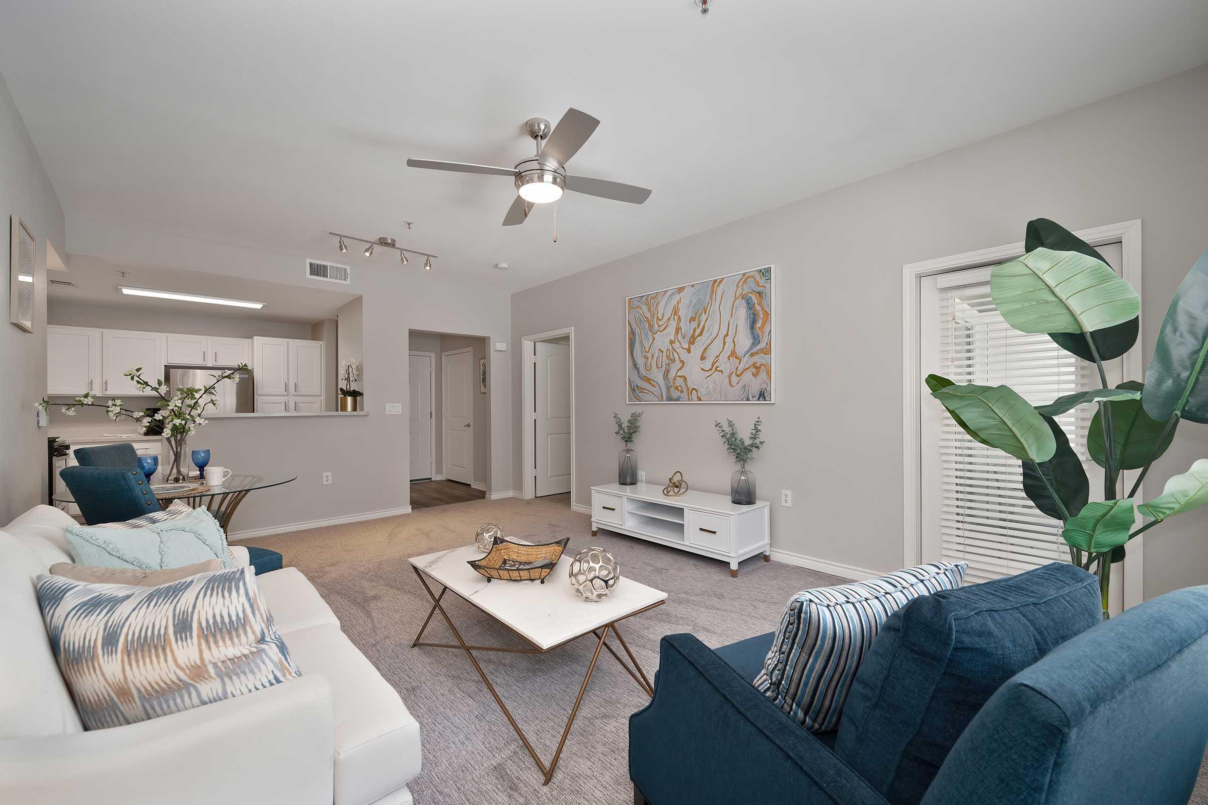 A modern living room featuring a white sofa and a blue armchair, with a coffee table in the center. The walls are light gray, adorned with a large abstract painting. There are potted plants and decorative items on the table. The space is well-lit with a ceiling fan and warm natural light coming through a window.