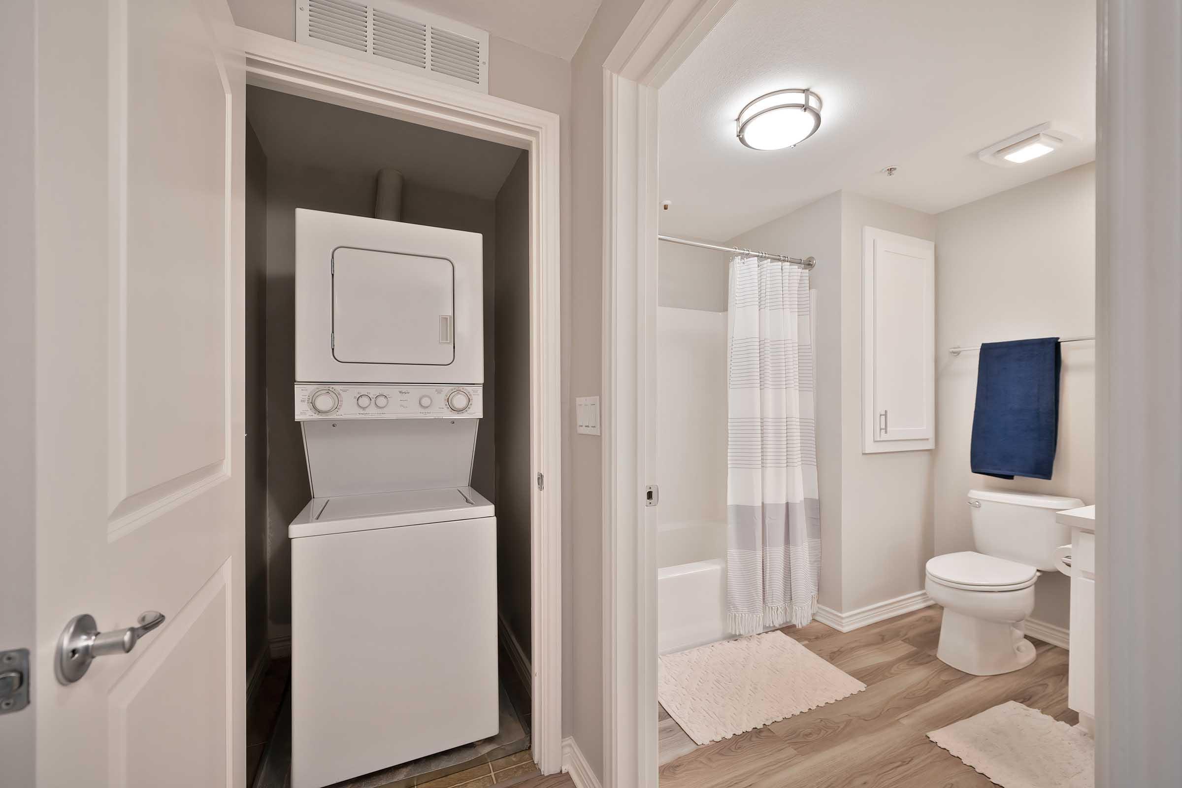 A clean, modern bathroom featuring a stacked washer and dryer unit in a corner, a bathtub with a shower curtain, a toilet, and white towels. Light wood flooring and neutral wall colors create a bright, inviting space.