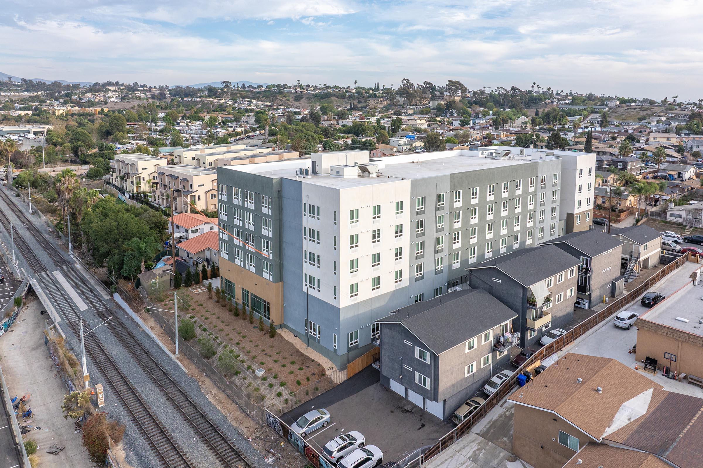 Aerial view of a modern multi-story building in an urban area, surrounded by smaller residential buildings and greenery. In the foreground, there are train tracks and parked cars, with hills in the background showcasing a suburban landscape. The sky is partly cloudy, contributing to a bright atmosphere.