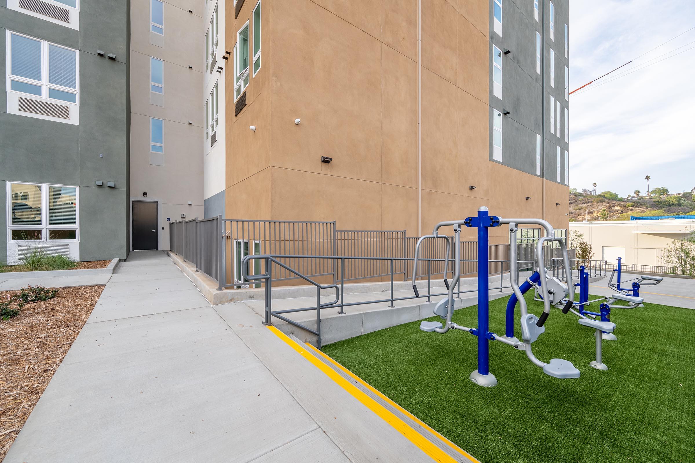 A modern apartment building with a playground area featuring exercise equipment on green turf. The building has a mix of gray and brown walls, and there is a ramp leading to the entrance. Surrounding landscaping includes mulch and small plants.