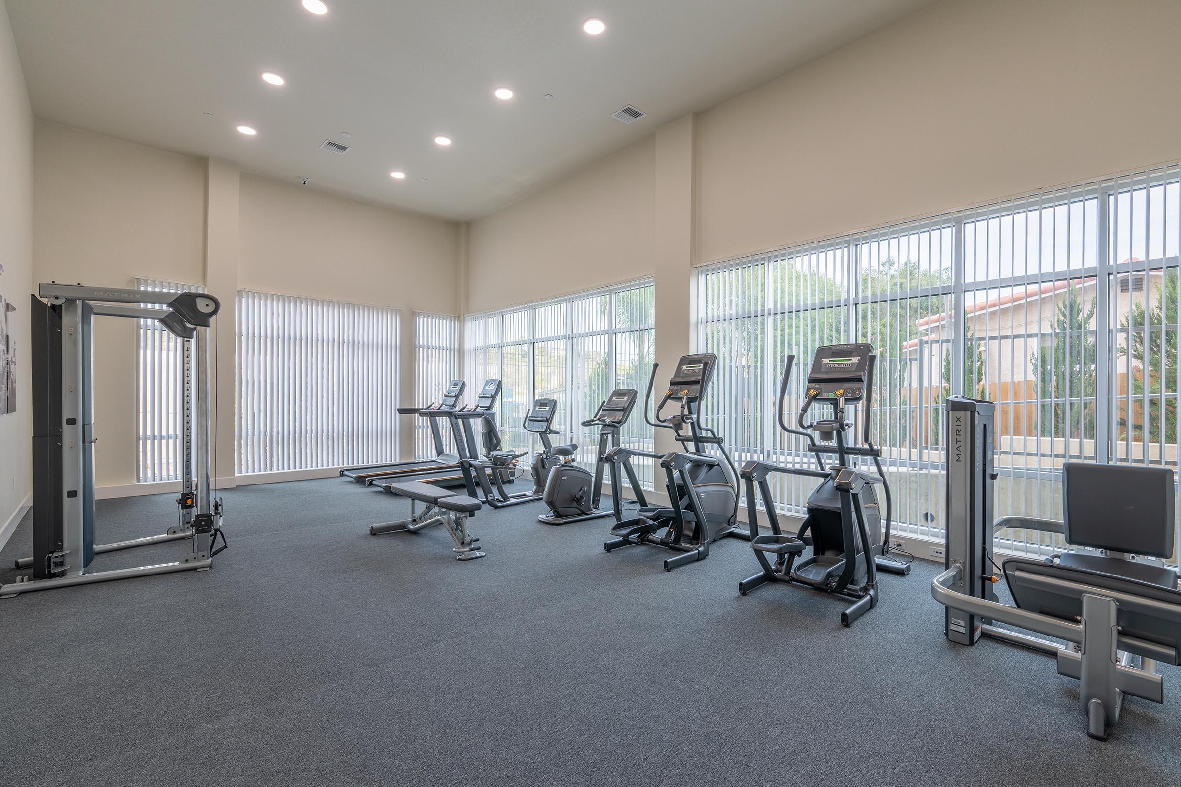 A spacious, well-lit gym interior featuring exercise equipment including treadmills, ellipticals, a cable machine, and a weight bench. The room has large windows with vertical blinds, allowing natural light to fill the space, and is equipped with gray carpeting.