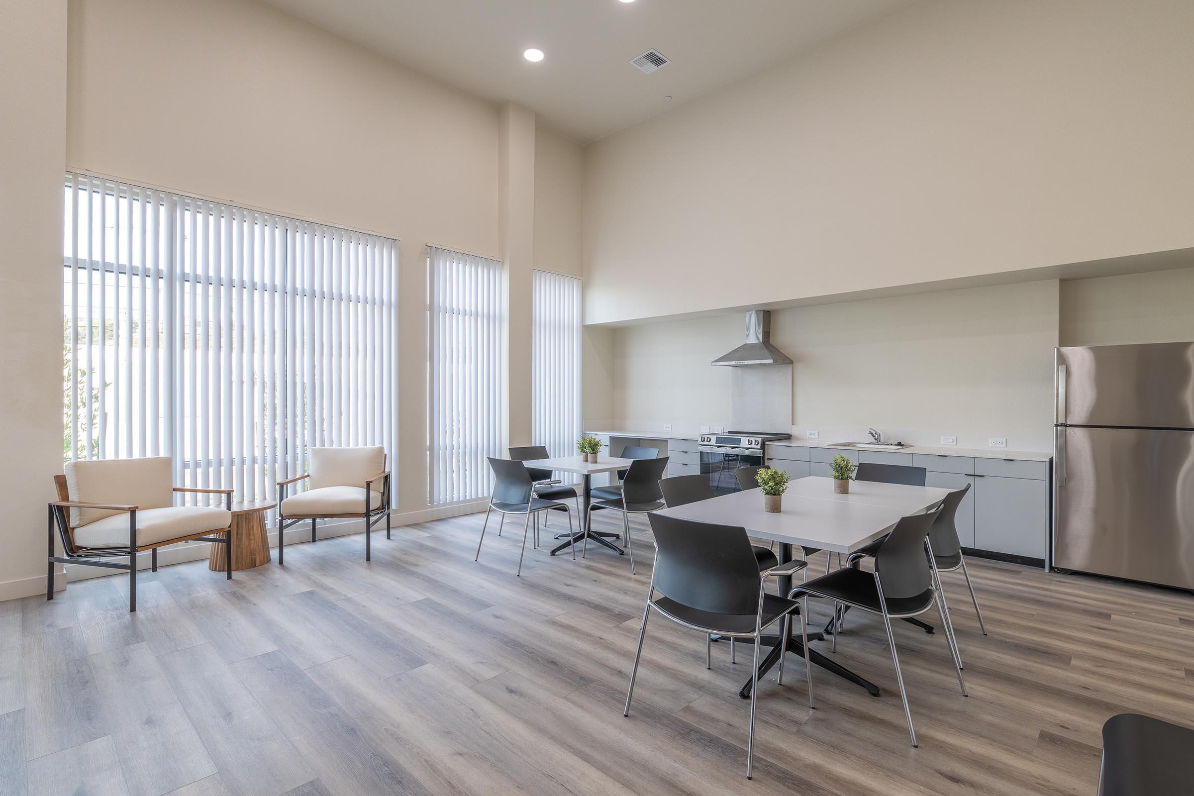 Modern kitchen and dining area featuring large windows with vertical blinds, light wooden flooring, and contemporary furniture. There are two armchairs to the left, several dining tables with chairs, and a kitchen with stainless steel appliances, including a stove and a refrigerator. Decorative plants are placed on tables.
