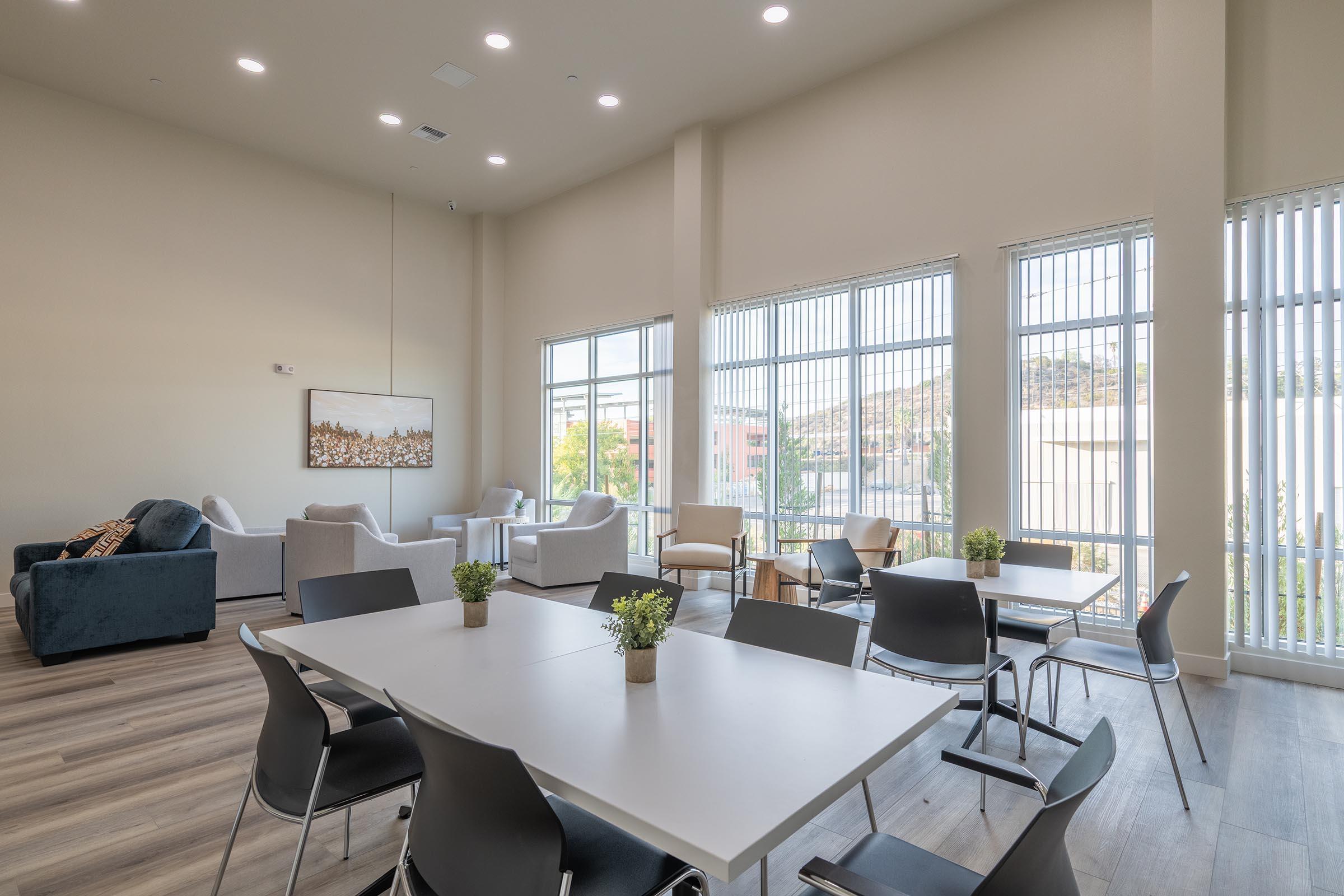 A bright, modern lounge area featuring several tables and chairs, a cozy seating arrangement with armchairs, and large windows that let in natural light. Potted plants are placed on the tables, and the decor is minimalistic, creating a welcoming atmosphere for socializing or relaxation.