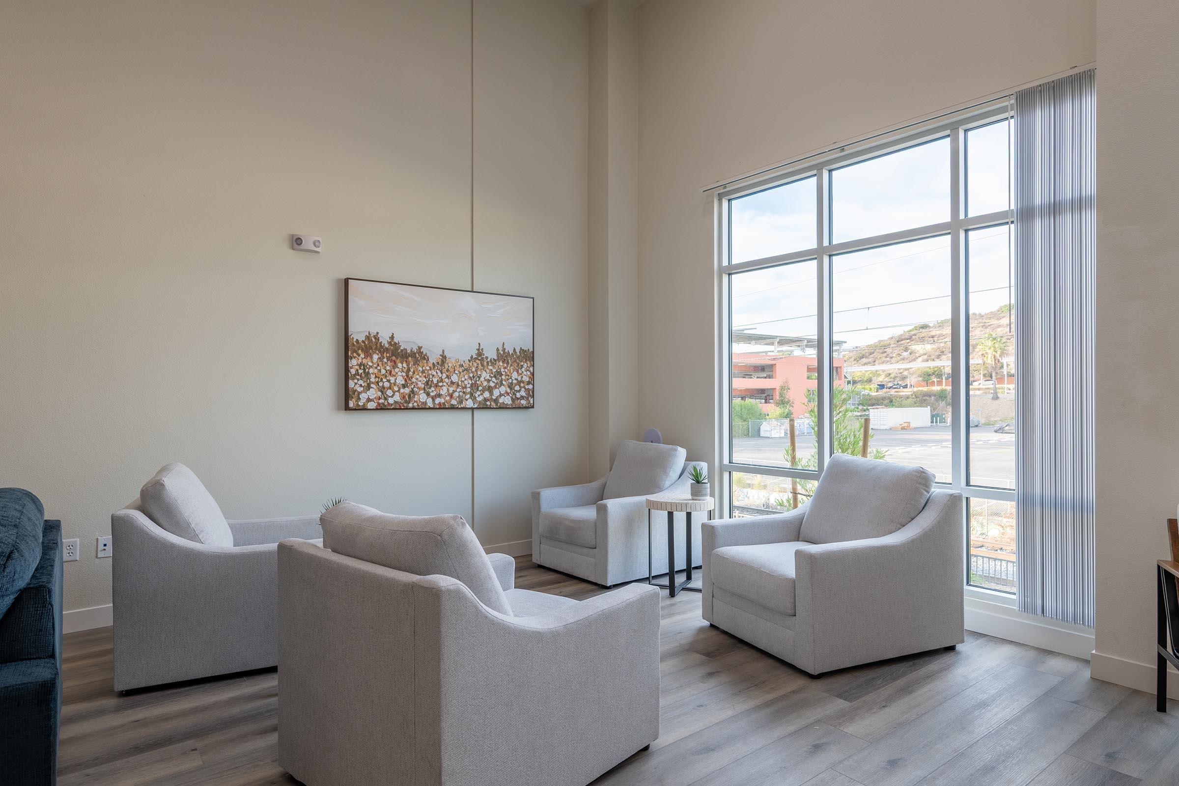 A modern living space featuring two light gray armchairs and a small side table. A piece of artwork is displayed on the wall, and large windows allow natural light to fill the room. The flooring is wooden, and there is a green couch visible in the corner. The overall atmosphere is bright and inviting.