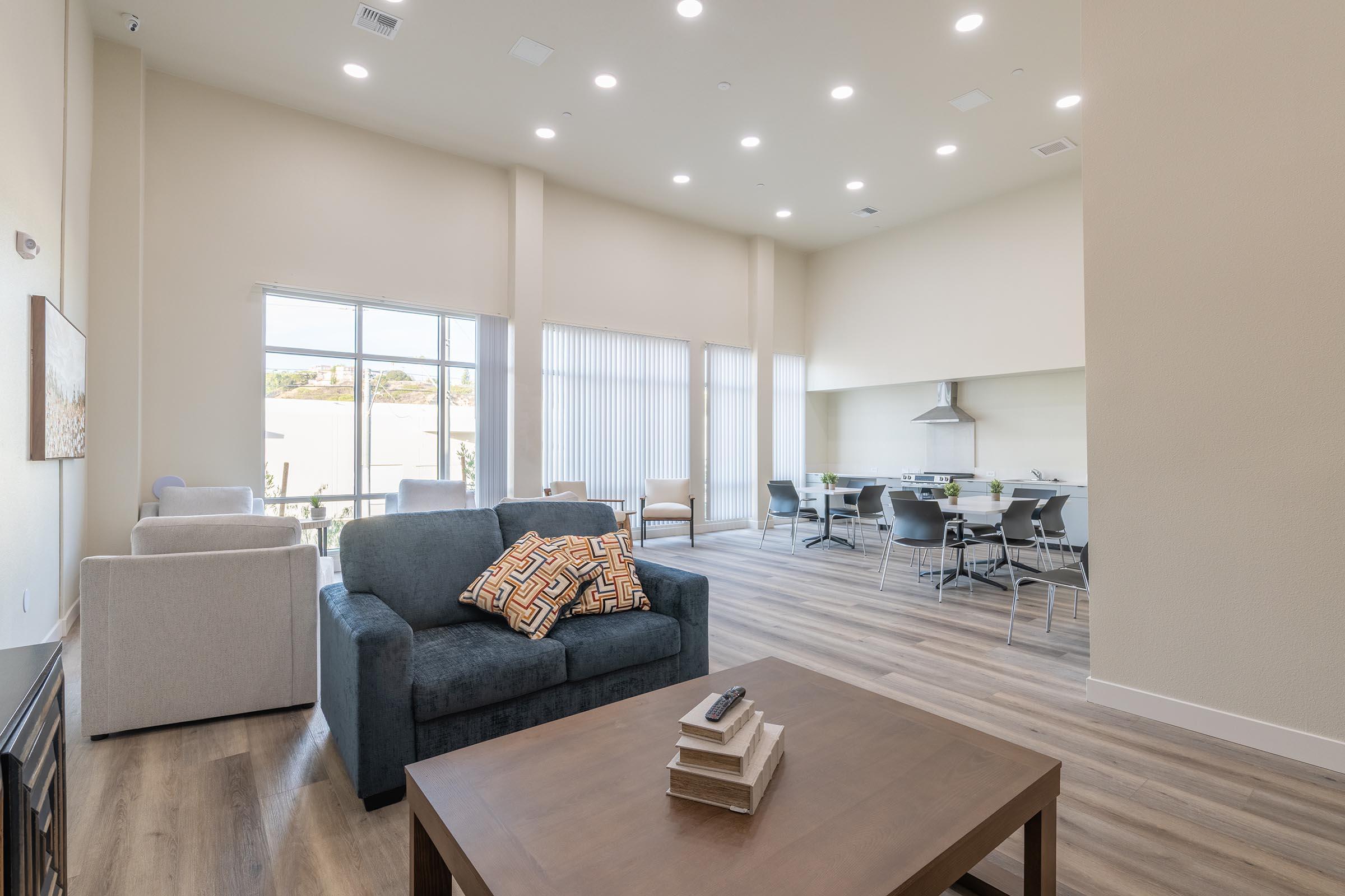 Modern lounge area featuring a blue sofa with a patterned pillow, light-colored armchairs, and a wooden coffee table. Large windows allow natural light to fill the space, which includes a dining area with a table and chairs. The floor has a wood-like finish, creating a warm and inviting atmosphere.