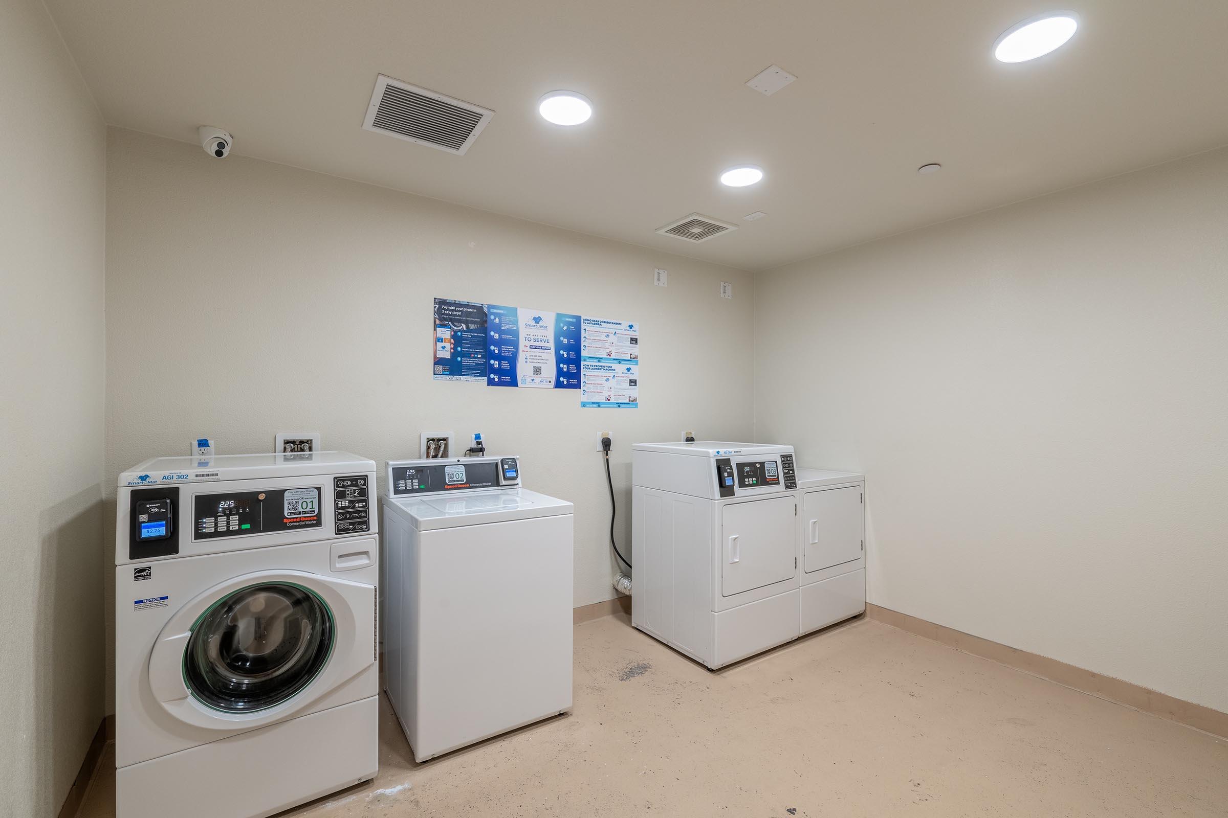 A clean, well-lit laundry room featuring two washing machines and one dryer. The machines are white and neatly arranged against a beige wall. There are informational posters on the wall and a ceiling-mounted camera for security. The floor appears polished and free of clutter.