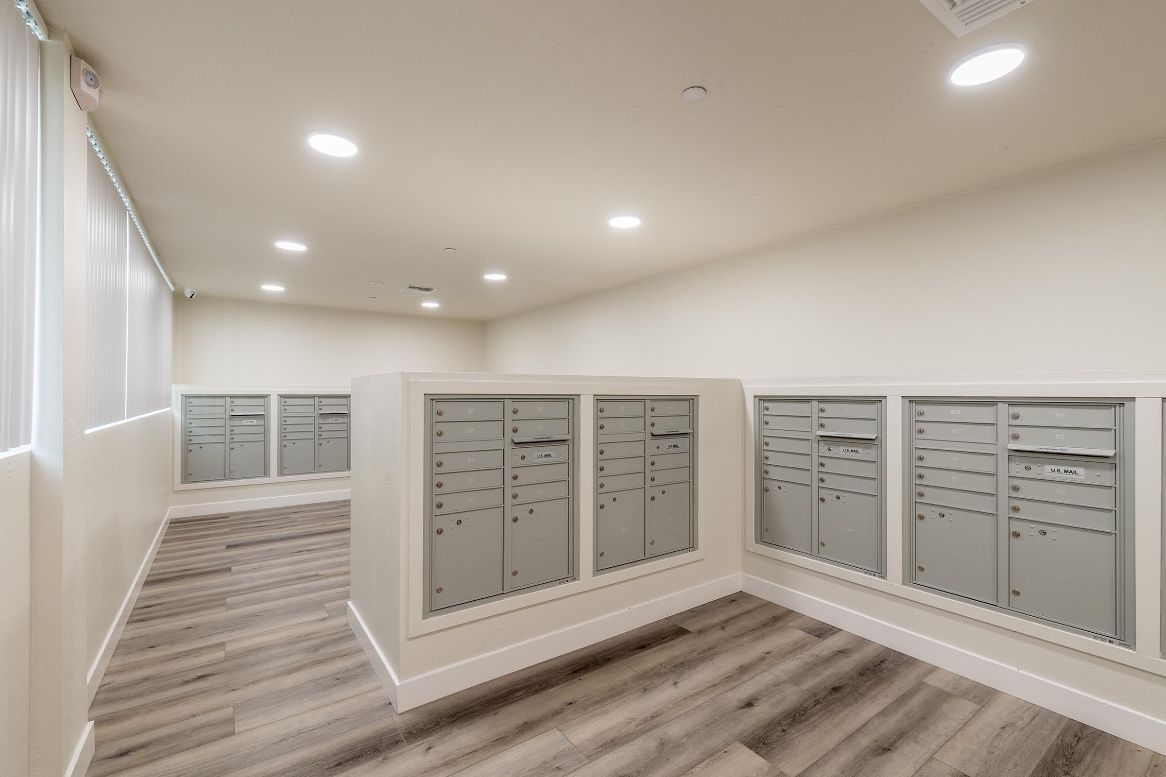 Interior view of a modern mailroom featuring multiple gray mailboxes arranged in two sections. The room has neutral-colored walls, wooden flooring, and bright overhead lighting. Large windows provide natural light, and the space appears clean and organized, ideal for residents to collect their mail.