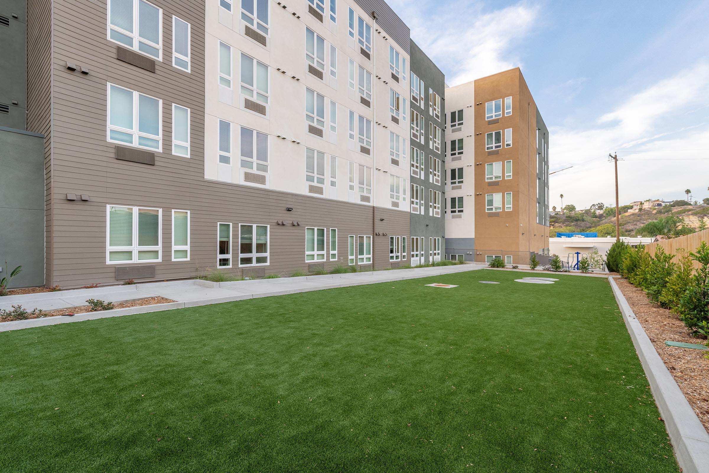 View of a modern multi-story residential building with large windows, surrounded by a well-maintained green lawn. The architecture features a mix of textures and colors, with concrete and wood-like finishes. The sky is partly cloudy, adding a bright ambiance to the outdoor space.
