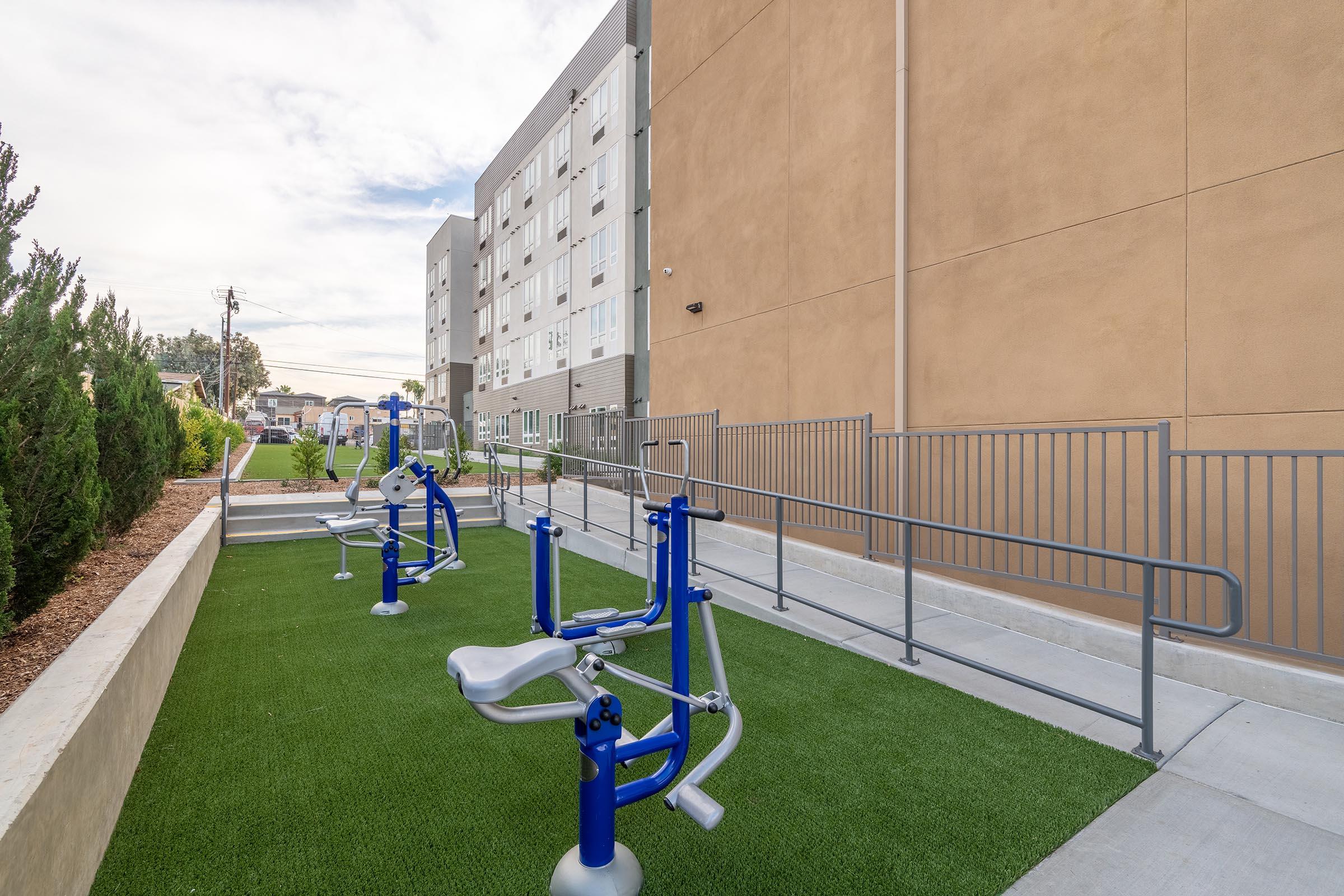 Outdoor fitness area with exercise equipment on synthetic green turf, adjacent to a modern building. The space features accessible ramps and is surrounded by landscaped shrubs and trees, creating a welcoming environment for physical activity.