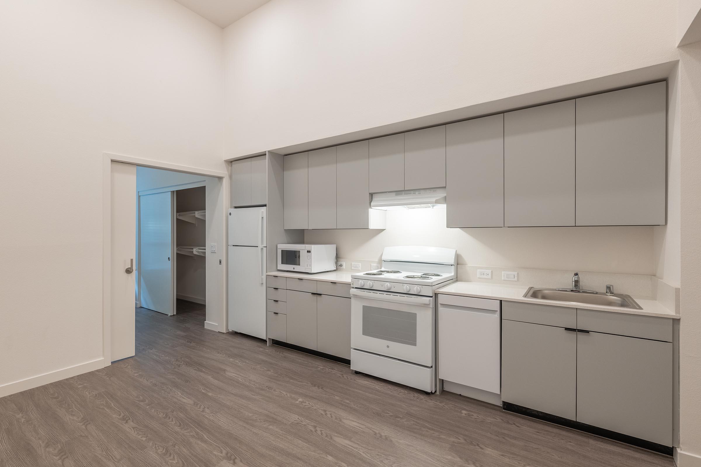 Modern kitchen with gray cabinets, white appliances including a stove, microwave, and refrigerator. A sink is situated next to the stove. The flooring is a light wood. An open doorway leads to another room, enhancing the spacious feel of the area with high ceilings and ample natural light.