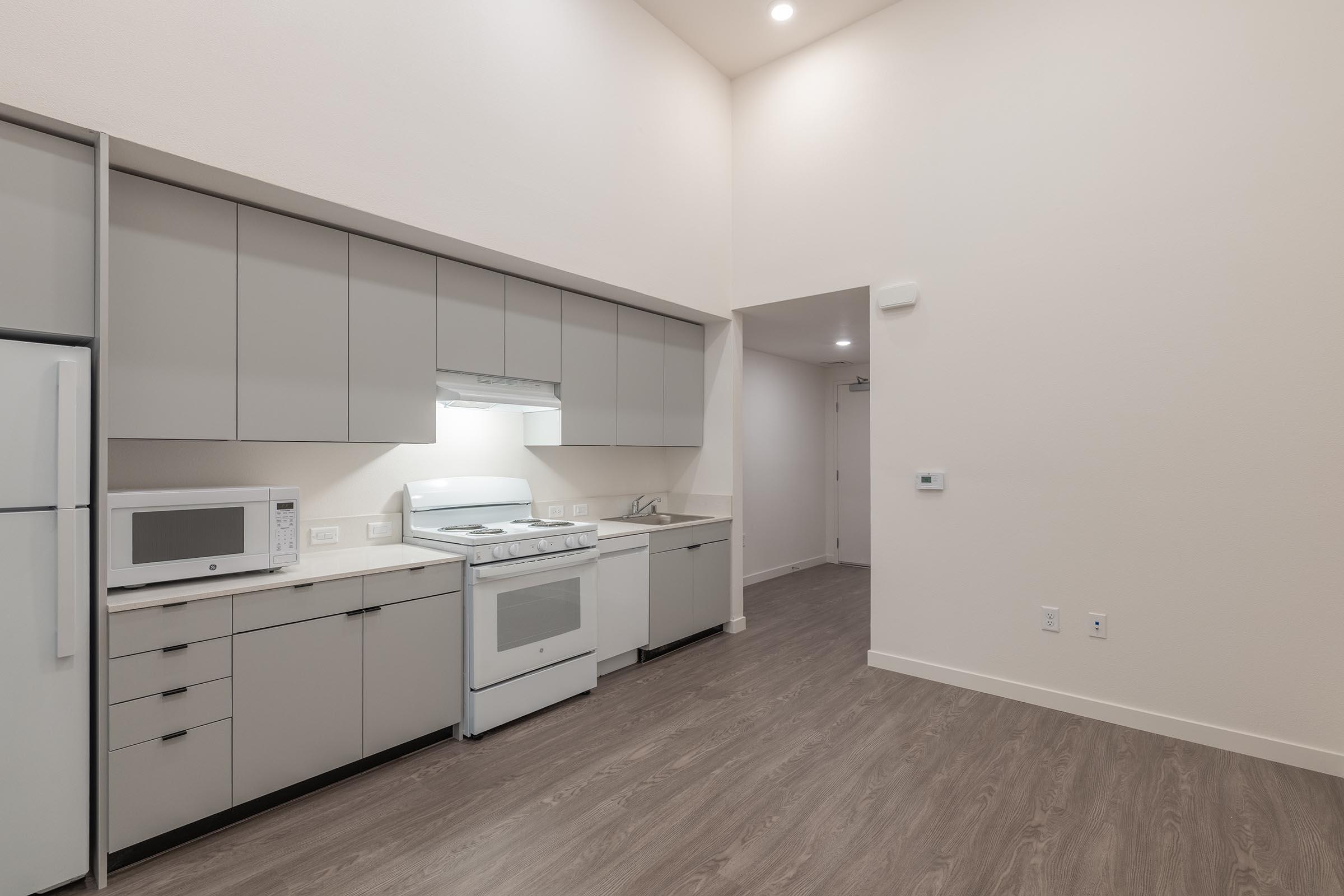 A modern kitchen featuring sleek gray cabinetry, a white stove, a microwave, and a refrigerator. The space has wood-like flooring and bright lighting from the high ceiling. An entrance to another room is visible in the background.