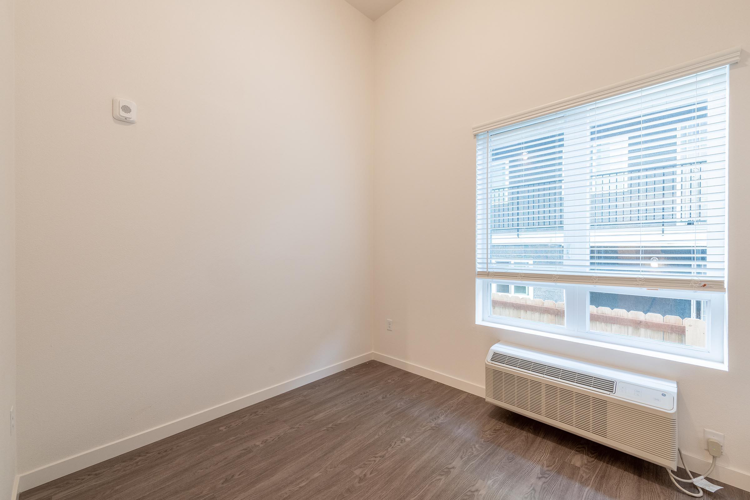 Empty room with white walls, a window with blinds, and an air conditioning unit beneath the window. The floor is finished with dark wood, and there is natural light coming through the window. The overall space is minimalistic and suitable for various purposes.