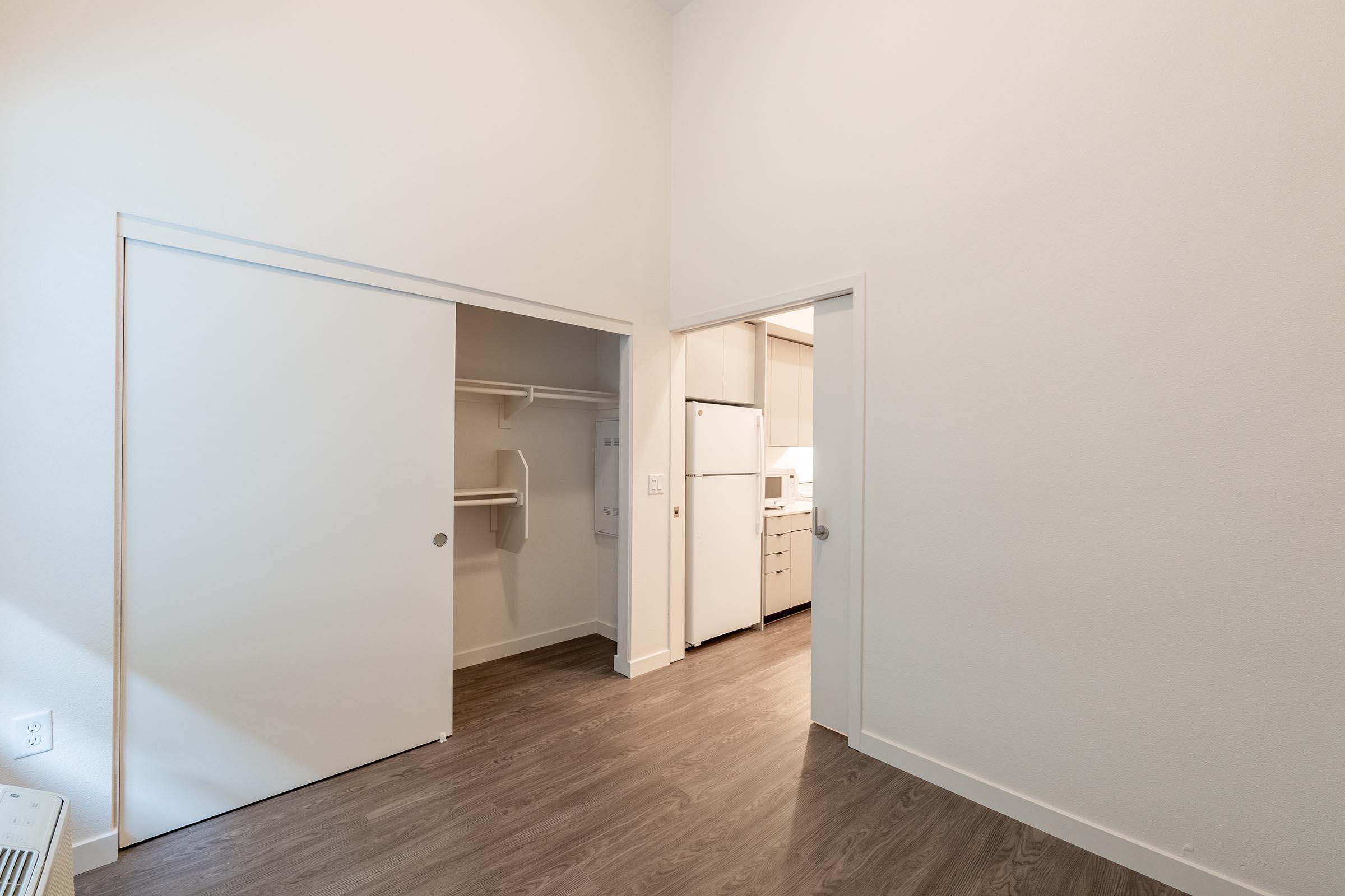 A well-lit, spacious room featuring two white closet doors on the left and an open doorway leading to a modern kitchen area on the right. The flooring is a light wood finish, and the walls are painted a soft white, creating a clean and inviting atmosphere.