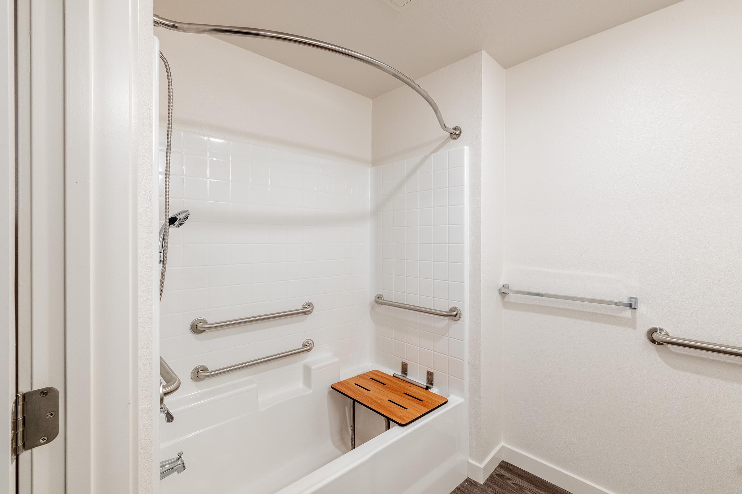 Bathroom featuring a bathtub with a wooden seat, grab bars on the walls for safety, and white tiled walls. The space is well-lit with a clean, modern design, emphasizing accessibility and functionality.