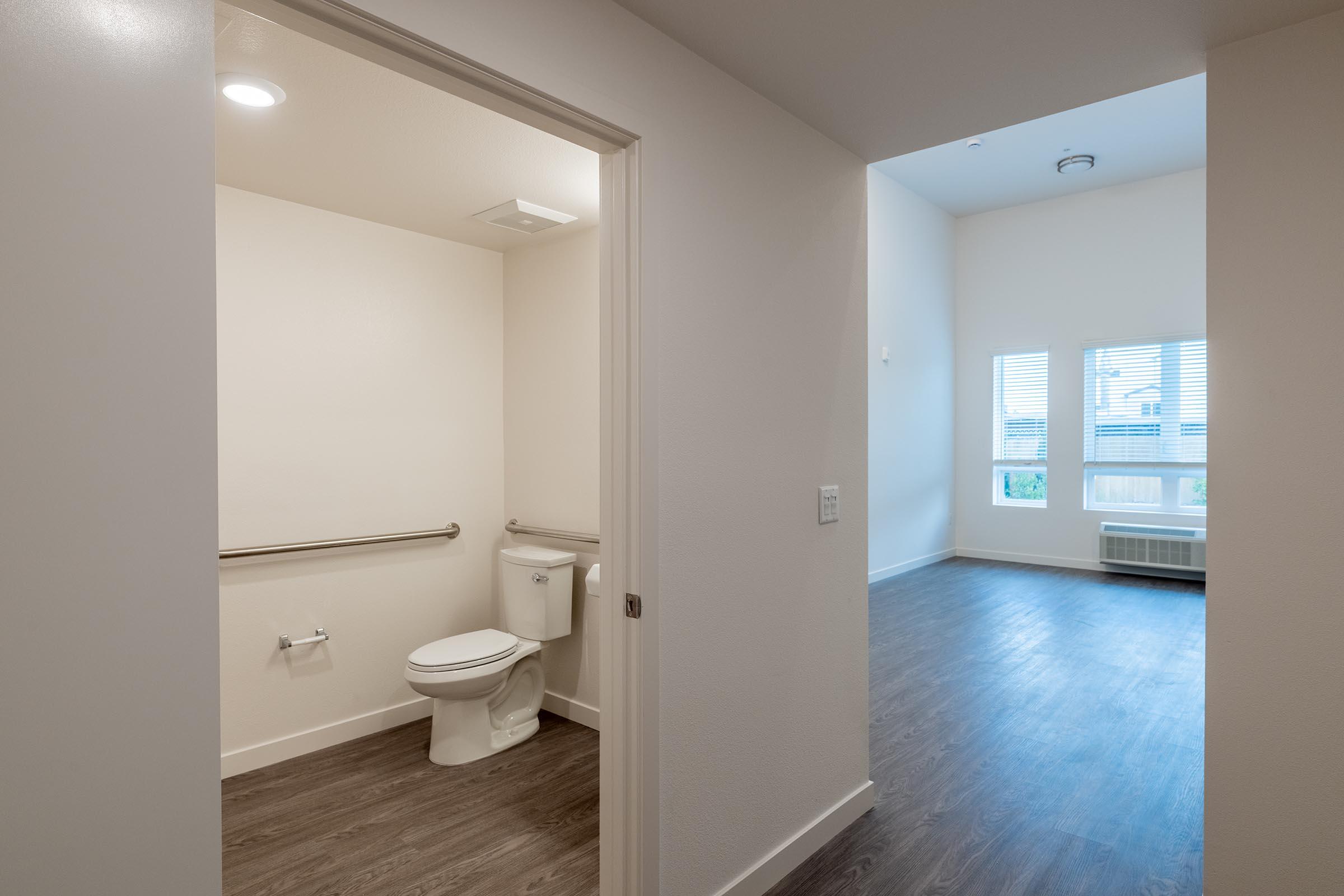 A view from an interior doorway showing a clean, modern bathroom on the left with a toilet and grab bar. On the right, a spacious, bright living area with multiple windows and light-colored walls. The flooring appears to be laminate wood, creating a contemporary aesthetic.