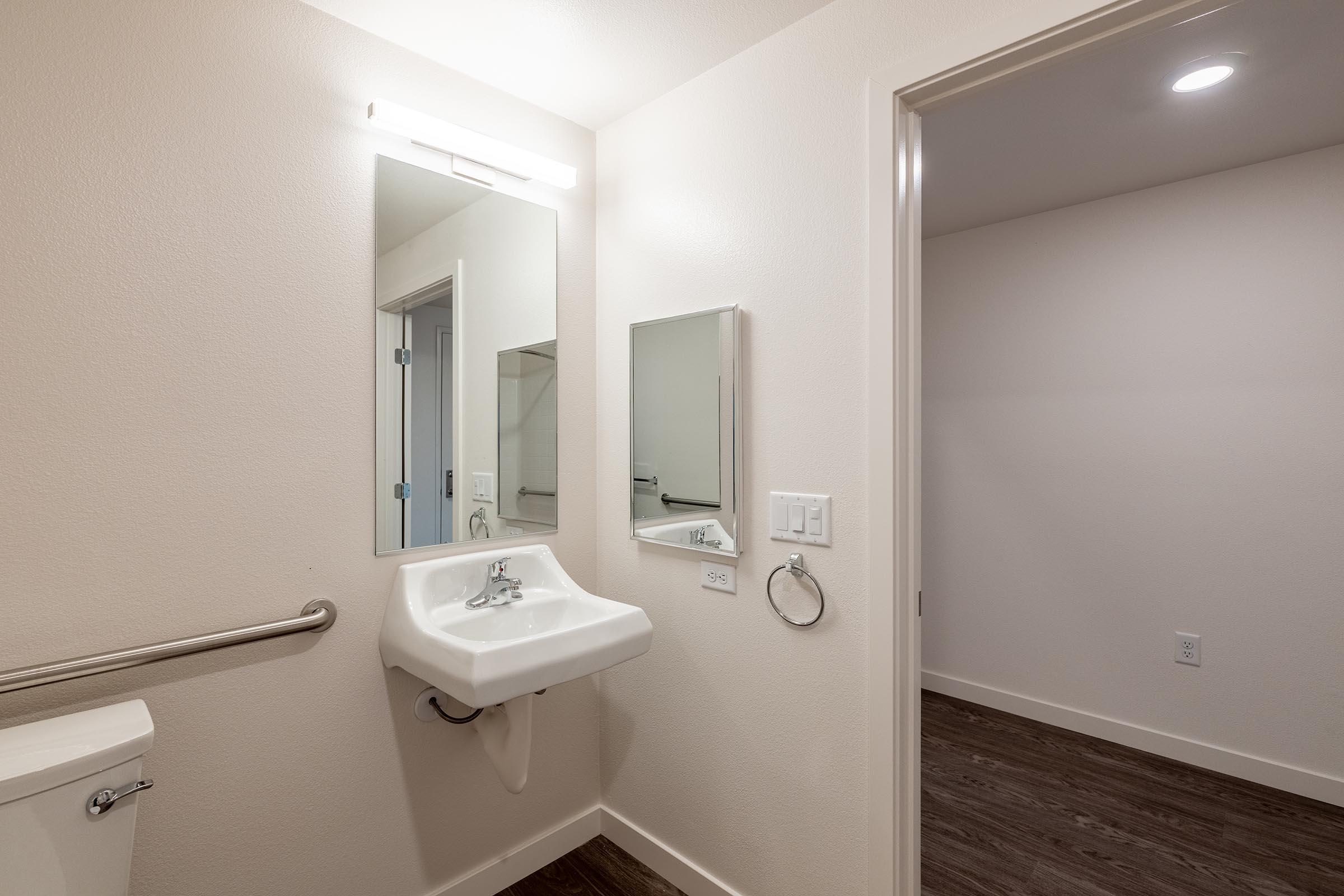 A modern bathroom featuring a white sink mounted on the wall, a large mirror above it, and a toilet to the left. The walls are painted in a light color, and the floor has dark wood-like tiles. A door leads to another room, and there are grab bars for accessibility near the sink.