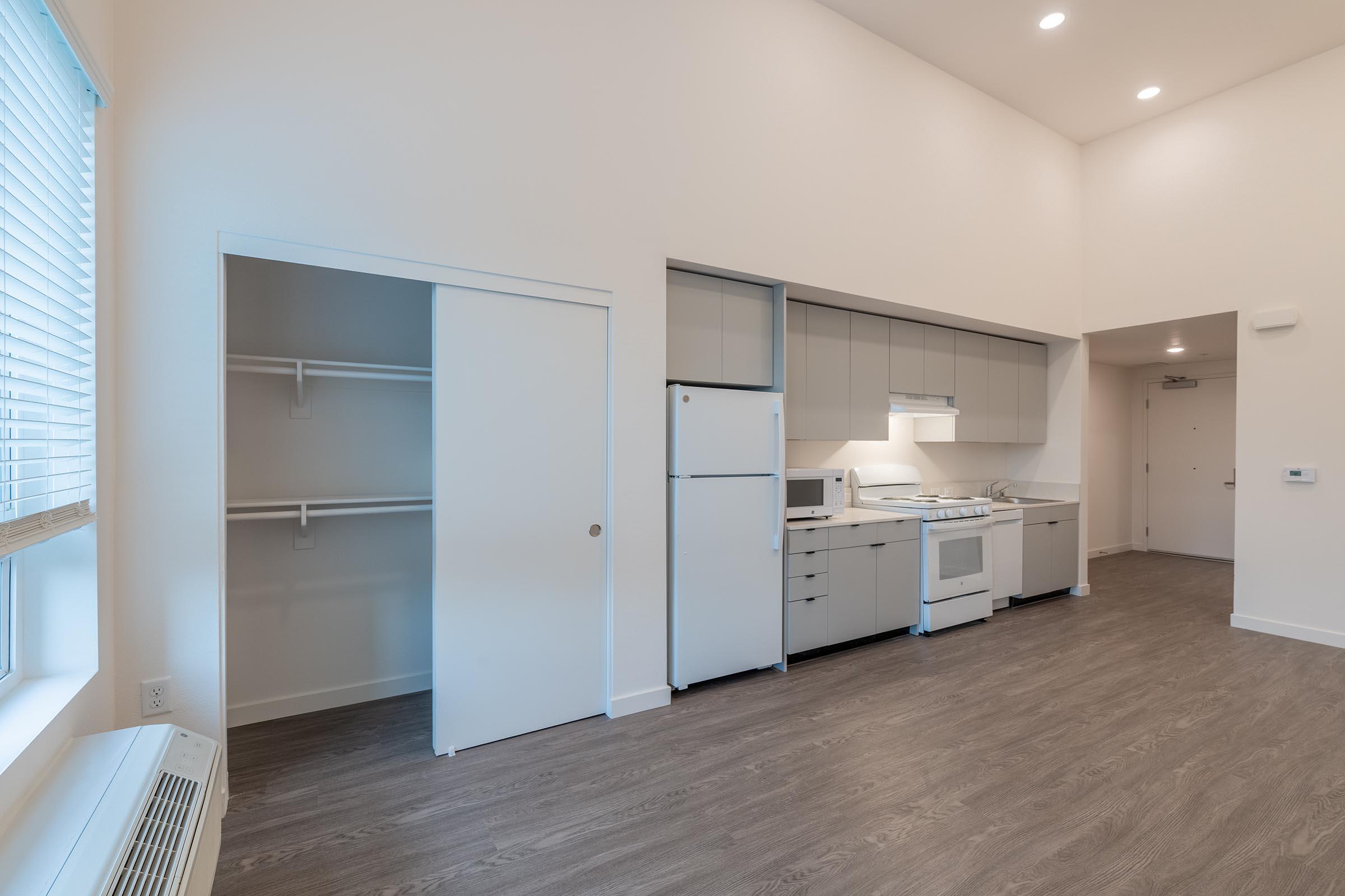 A modern kitchen and living space featuring a white refrigerator, gas stove with oven, microwave, and light-colored cabinetry. There's a closet with sliding doors on the left and a spacious layout with high ceilings and natural light from a window. The flooring is a warm wood tone.