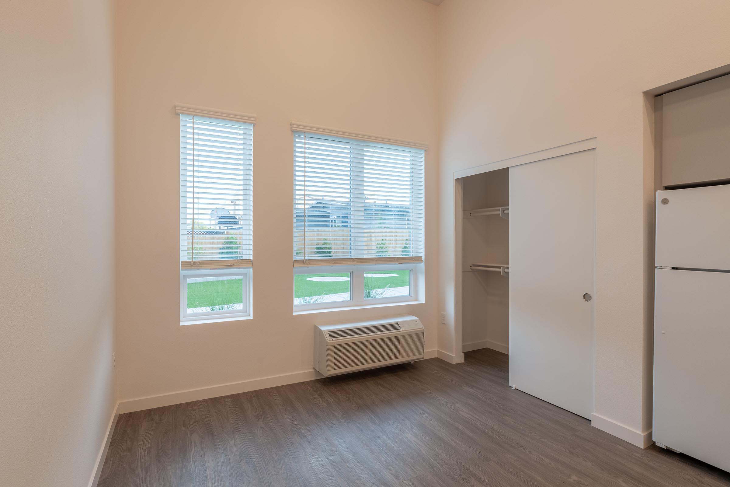 A bright, minimalist room featuring light-colored walls and wooden flooring. Two large windows with blinds provide natural light, and there is a small closet on the left. An air conditioning unit is installed below the windows, and a white refrigerator is visible in the corner. The overall atmosphere is modern and inviting.