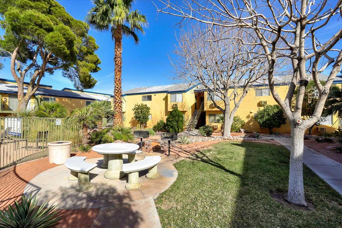 A landscaped courtyard featuring palm trees and bare trees, with a circular stone table and benches surrounded by greenery. Yellow buildings are in the background under a clear blue sky, creating a welcoming outdoor space for relaxation.