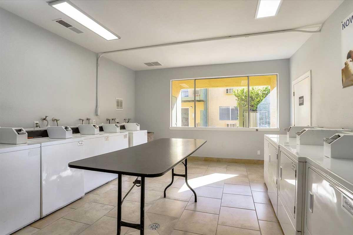 A clean, spacious laundry room with several washing machines lined against the wall. A central table is set in the middle, and there is a large window allowing natural light to fill the space. The walls are light-colored, creating an inviting atmosphere for doing laundry.