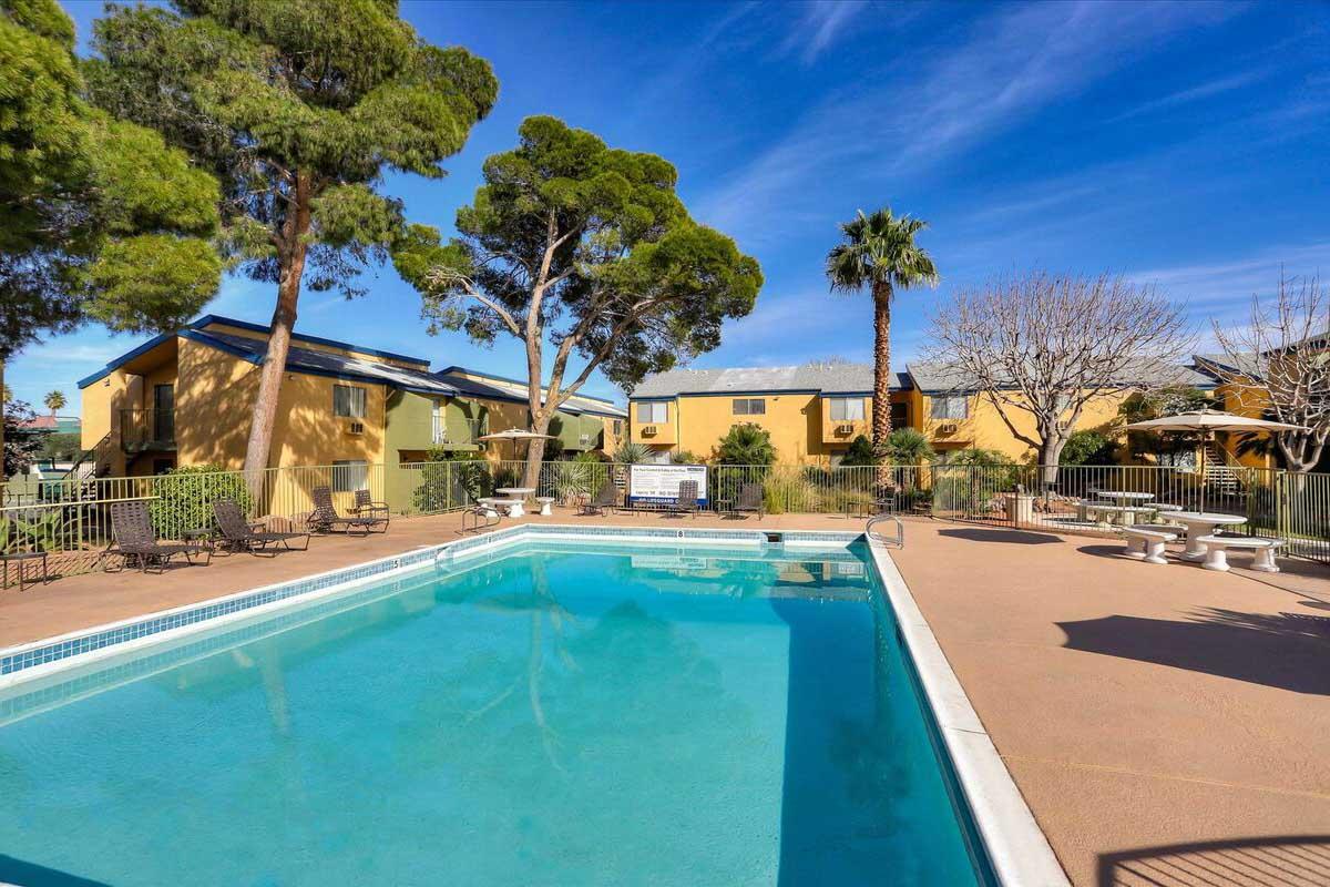 A clear swimming pool surrounded by trees in a residential area, with lounge chairs and tables nearby. There are two-story buildings in the background, featuring yellow exteriors and greenery. The sky is blue with a few clouds, creating a relaxing atmosphere.