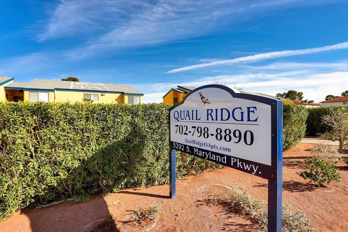 Sign for Quail Ridge located at 5292 S. Maryland Pkwy, featuring the phone number 702-798-8890 and website URL QuailRidgeVIPApts.com, surrounded by green shrubbery and a clear blue sky.