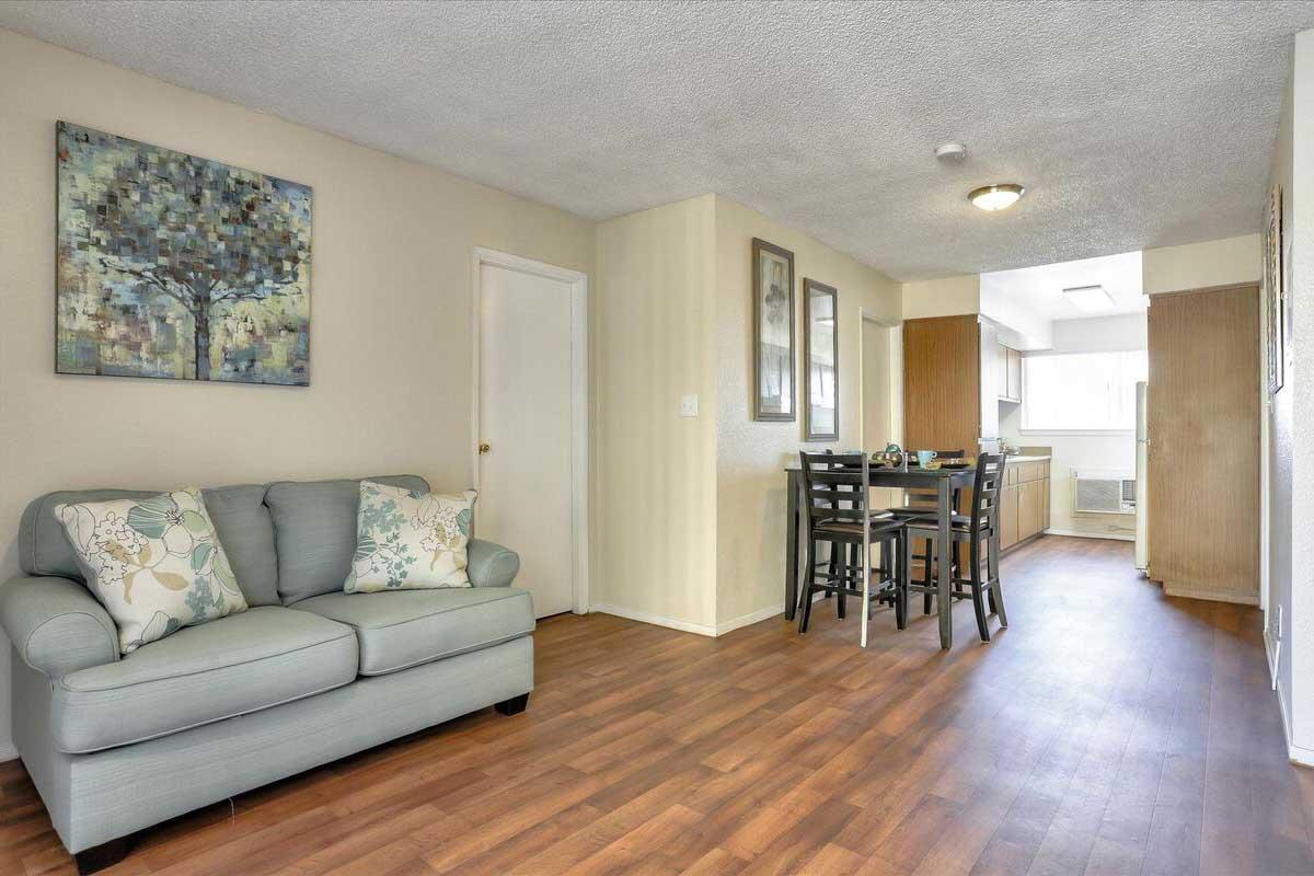 A cozy living space featuring a light blue sofa adorned with decorative pillows, a dining area with a table and chairs, and a hallway leading to a kitchen. The walls are painted in a soft beige, and there's a piece of art depicting a tree. The flooring is wood-like, creating a warm and inviting atmosphere.