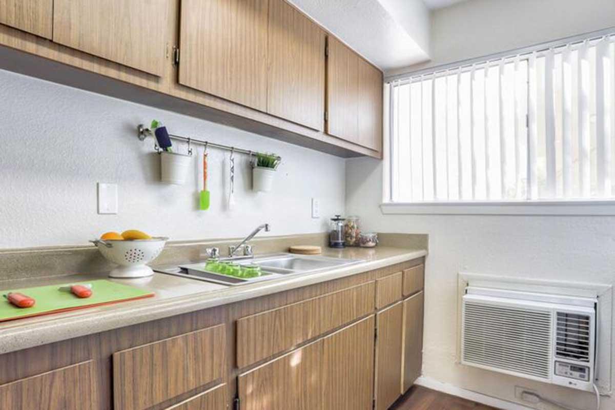 A modern kitchen featuring wooden cabinets, a double sink, and a light countertop. A cutting board with tomatoes sits on the counter, alongside a bowl of oranges. An air conditioning unit is installed below a window with blinds, allowing natural light into the space.