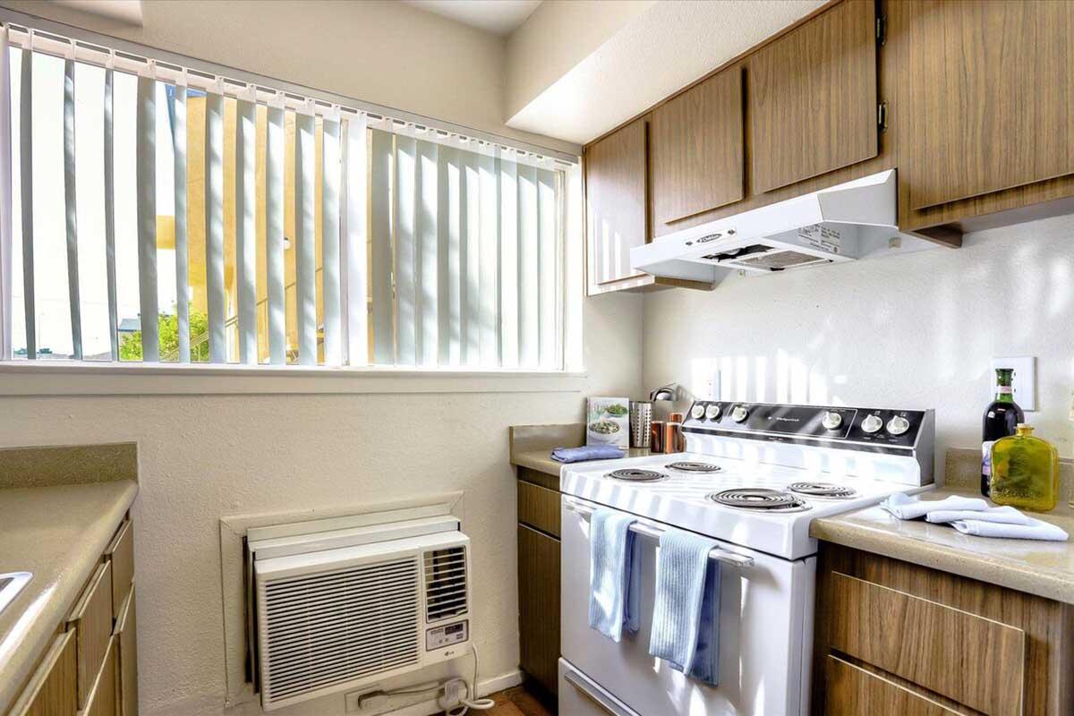 A bright kitchen with wooden cabinets, featuring a white stove and an exhaust hood. An air conditioning unit is installed below a window adorned with vertical blinds. The countertop holds a bottle of olive oil and kitchen accessories, creating a cozy and functional space.