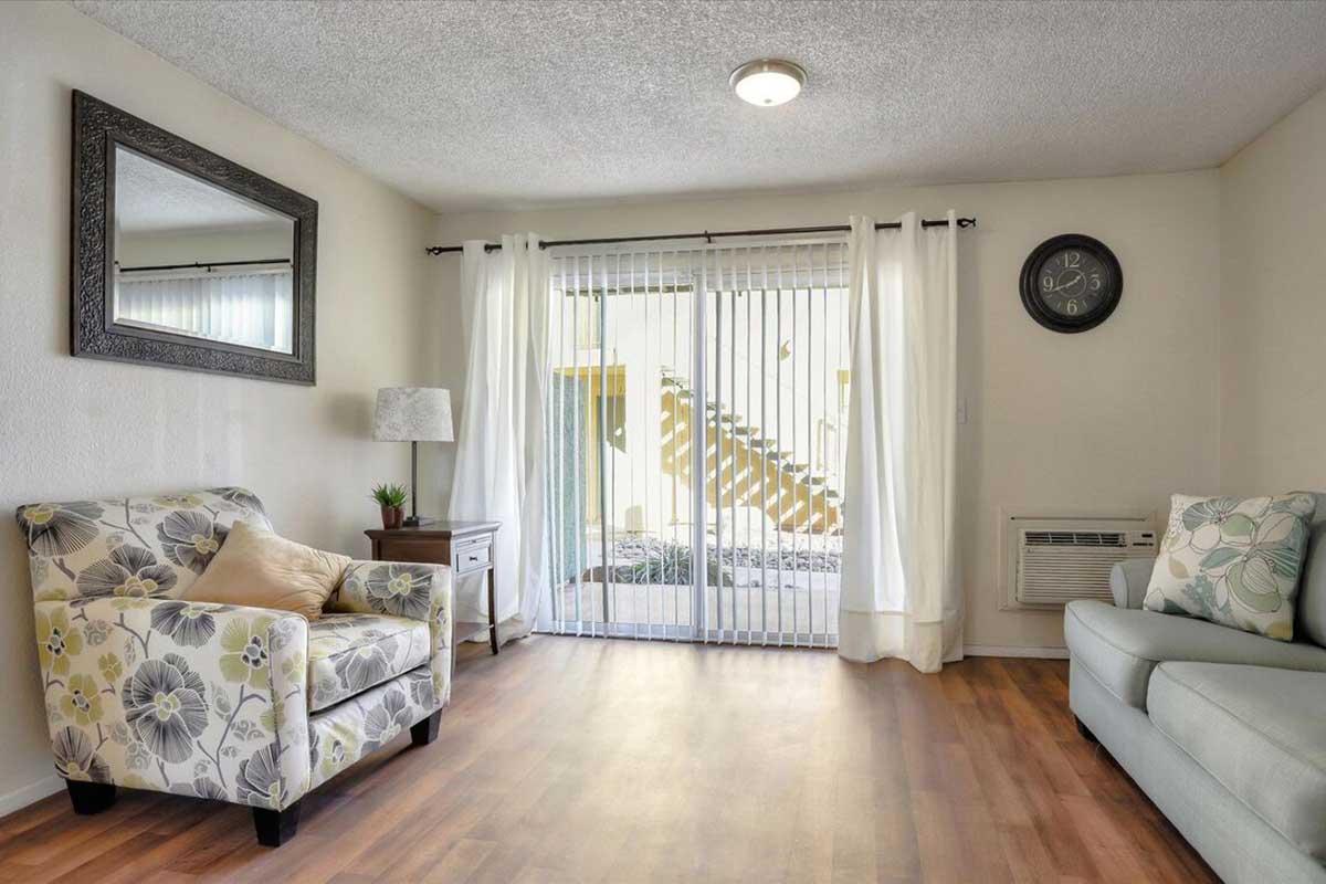 A cozy living room featuring two armchairs with floral patterns, a light gray sofa, and a wooden side table with a lamp. There is a large mirror on the wall, a clock, and sheer curtains allowing light to filter through. A view of stairs outside is visible through the sliding glass door.